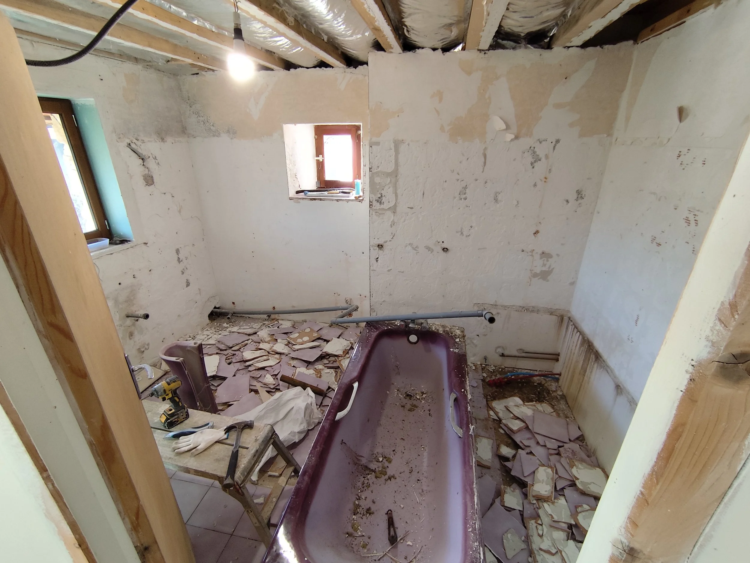 A room under renovation with debris on the floor, exposed ceiling beams with insulation, and unfinished walls. There is a small window on the wall with tools and materials nearby.