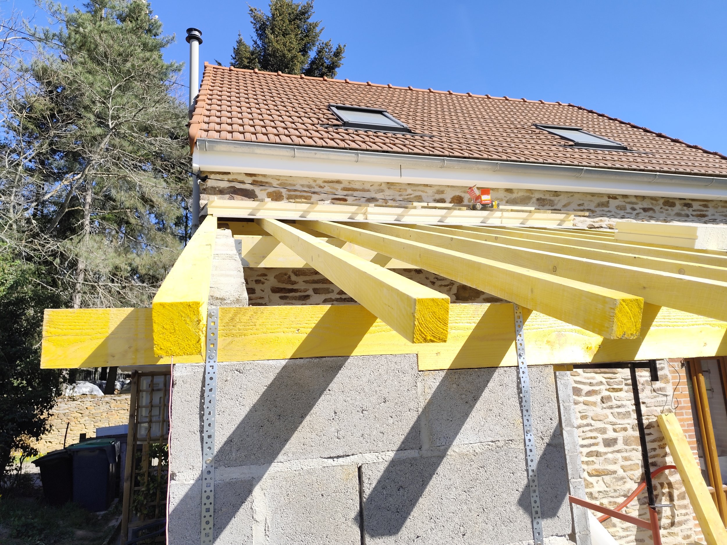 Construction scaffolding with yellow wooden beams in front of a house with stone and stucco exterior, two skylights on the roof, and trees in the background.