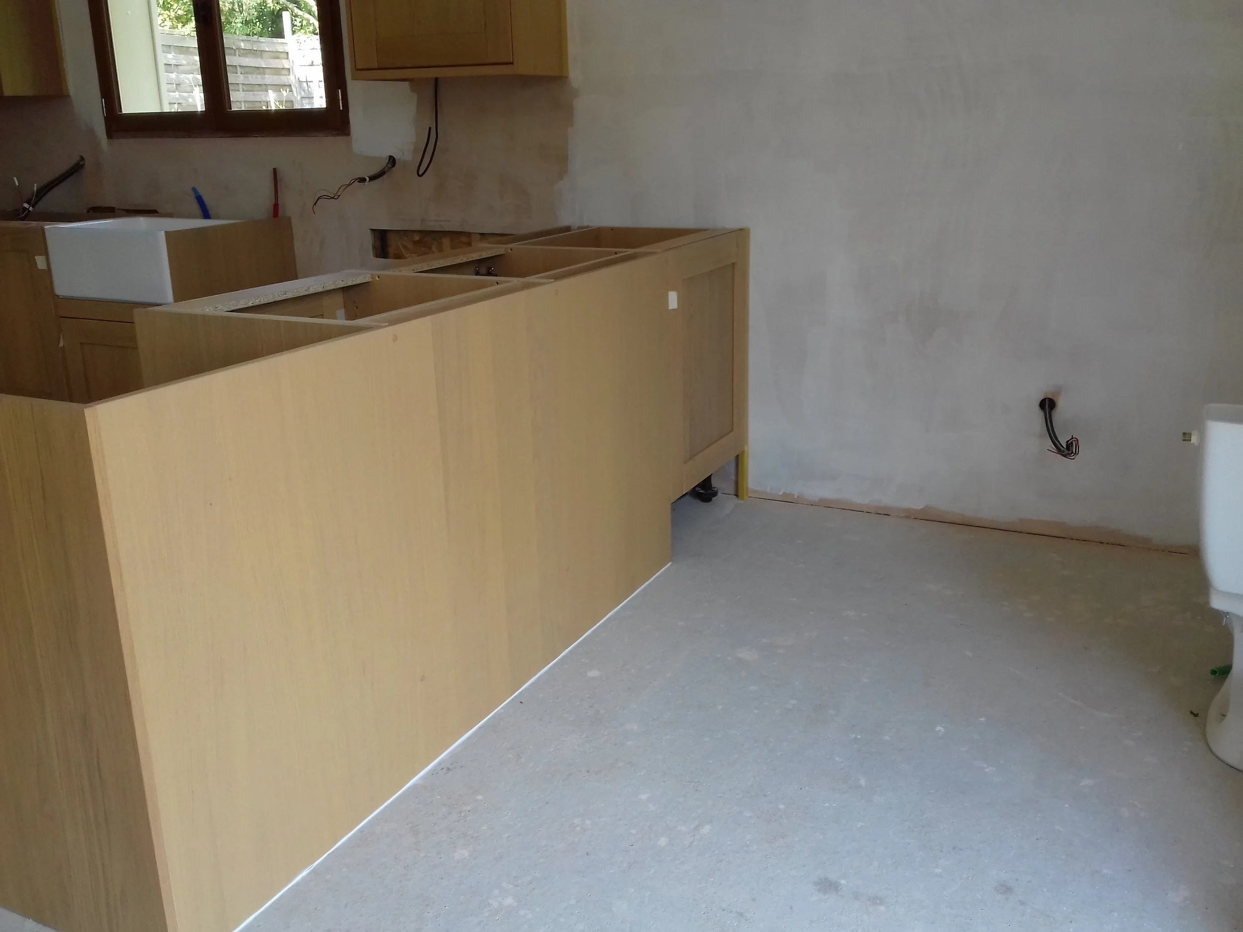 Inside a room under construction or renovation, with unfinished wooden cabinets and a single white sink. The walls are partially painted, and there are exposed electrical wires. A window and a small cabinet are visible near the ceiling.