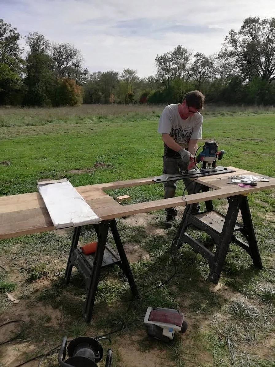 Person using a power tool on a piece of wood outdoors, with a large grassy field and trees in the background.