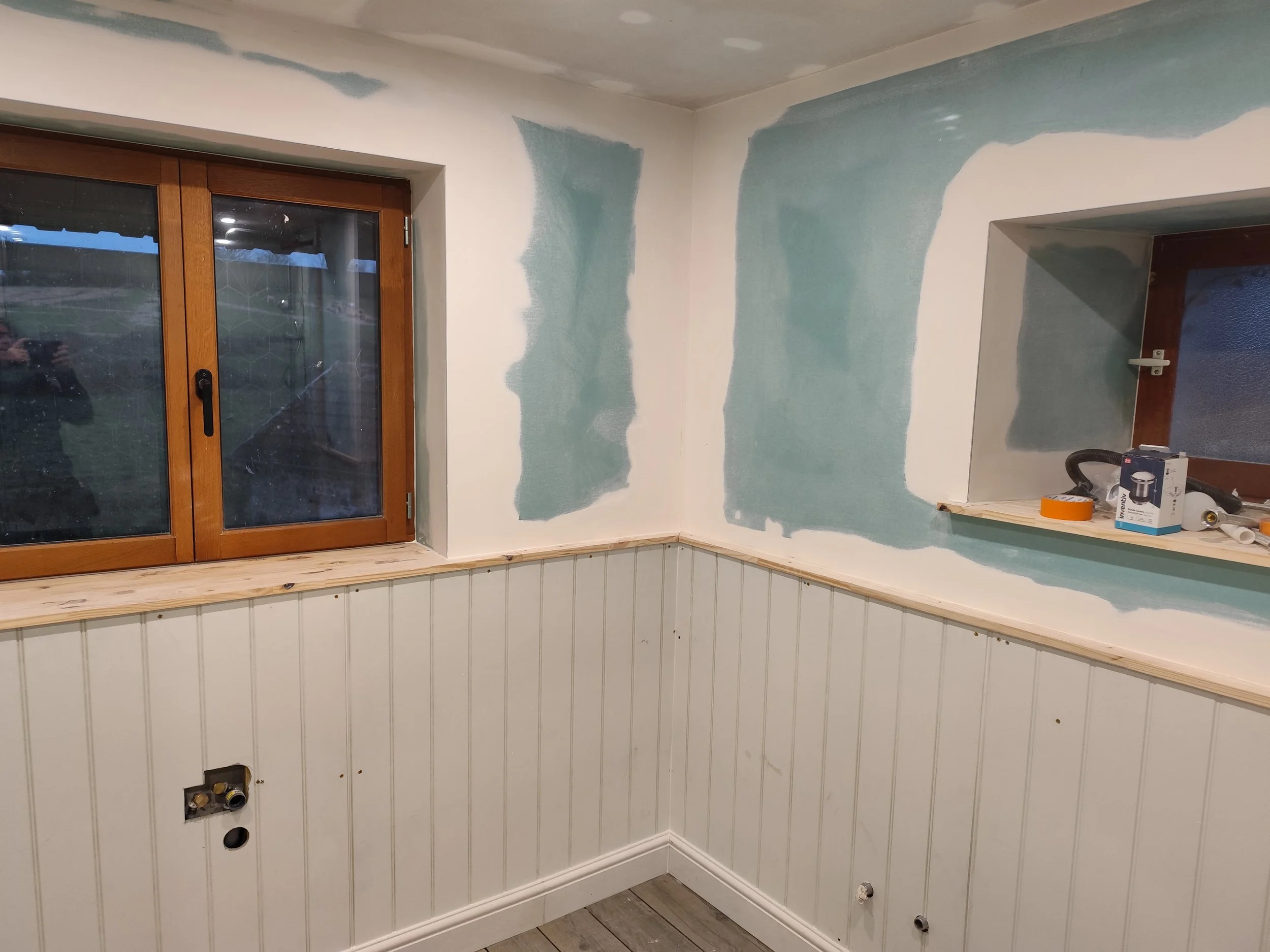 Indoor room undergoing renovation, walls partially painted with drywall and paint patches, with plumbing fixtures and tools visible.