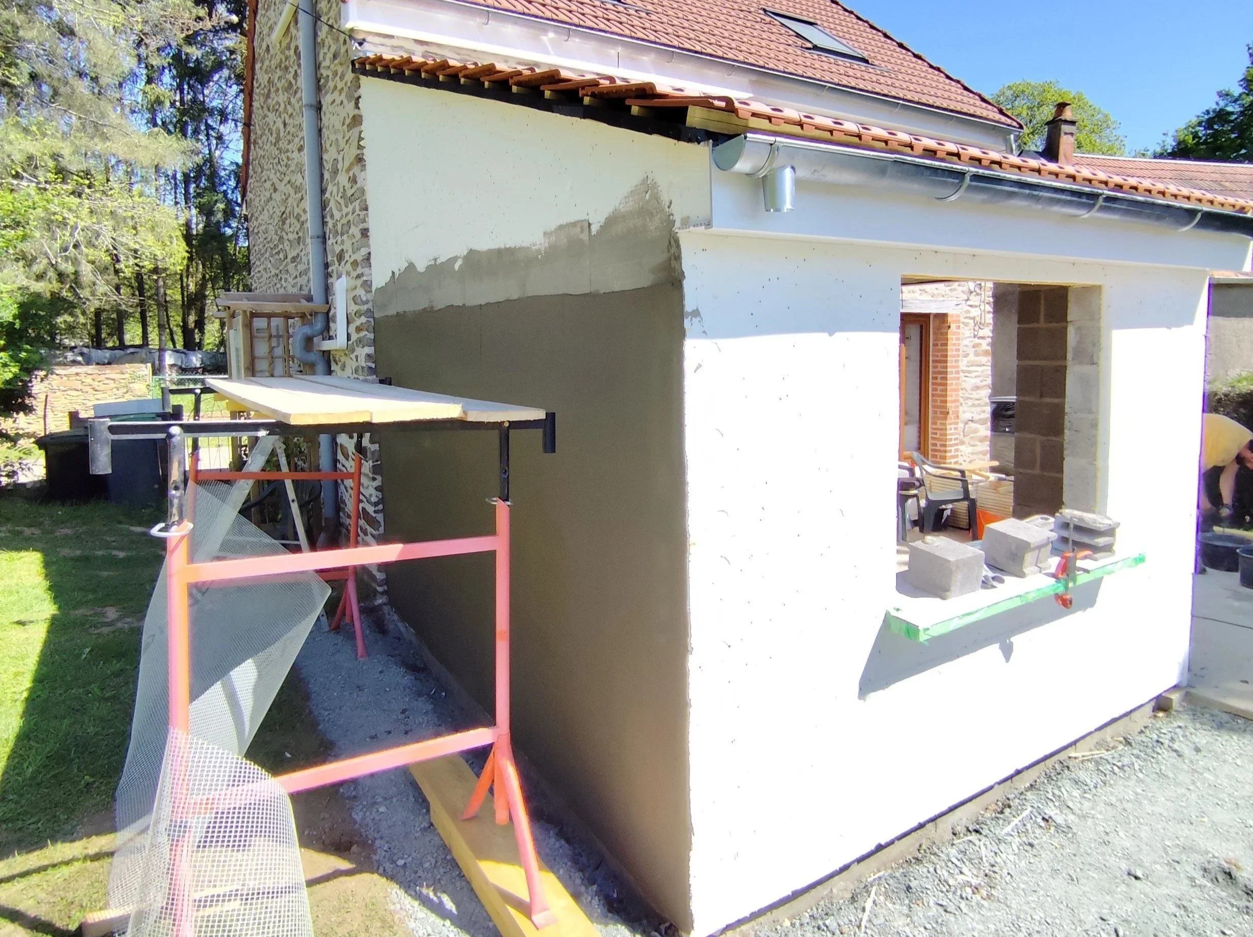 A house under construction with a partially finished exterior wall, construction tools, and materials around it, with trees and a clear blue sky in the background.