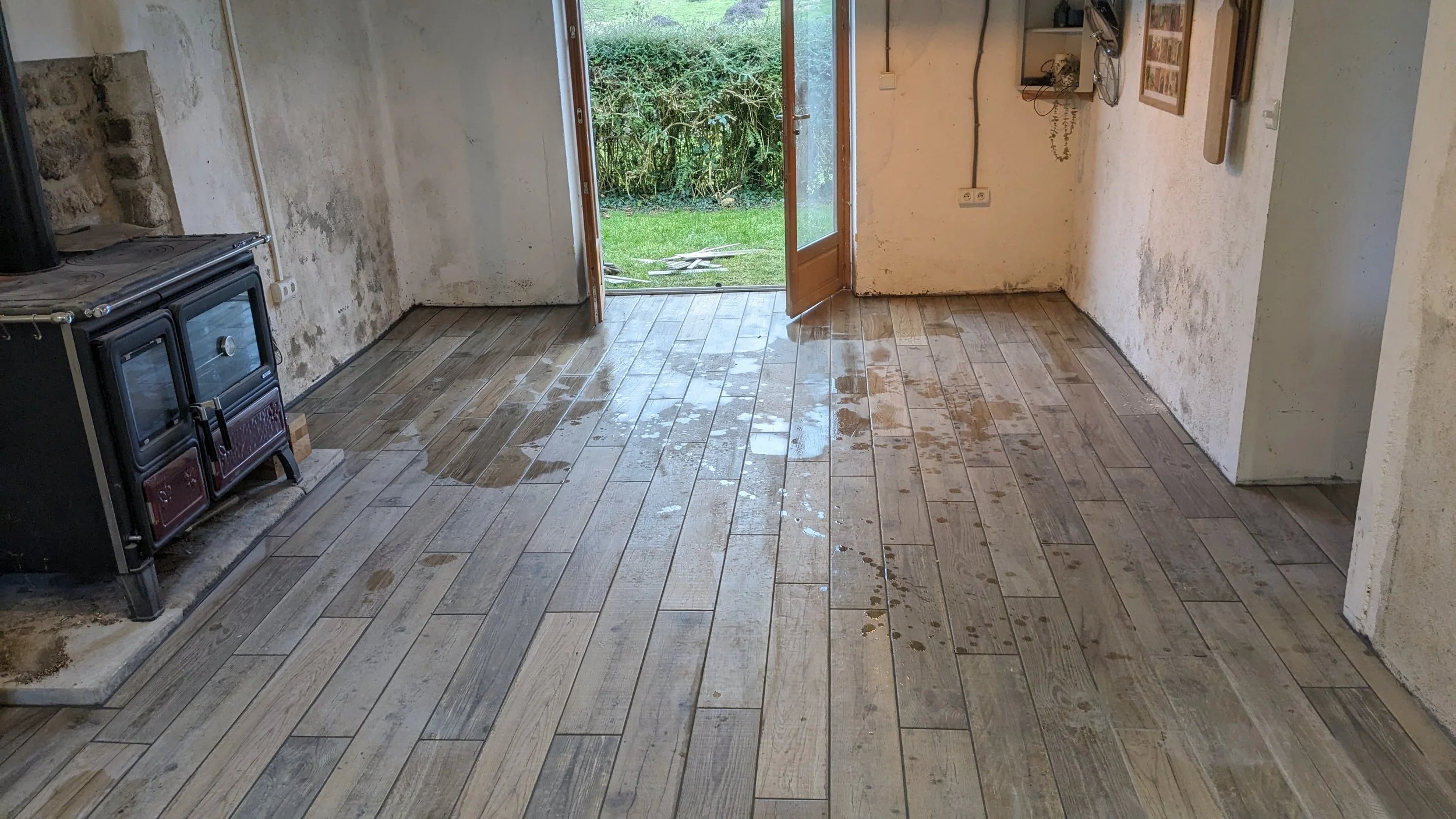 A room with wooden flooring that appears to have been recently mopped, with water on the surface. An open door leads outside to a grassy yard with bushes. There is an old stove on the left side and some framed pictures or artwork on the right wall.