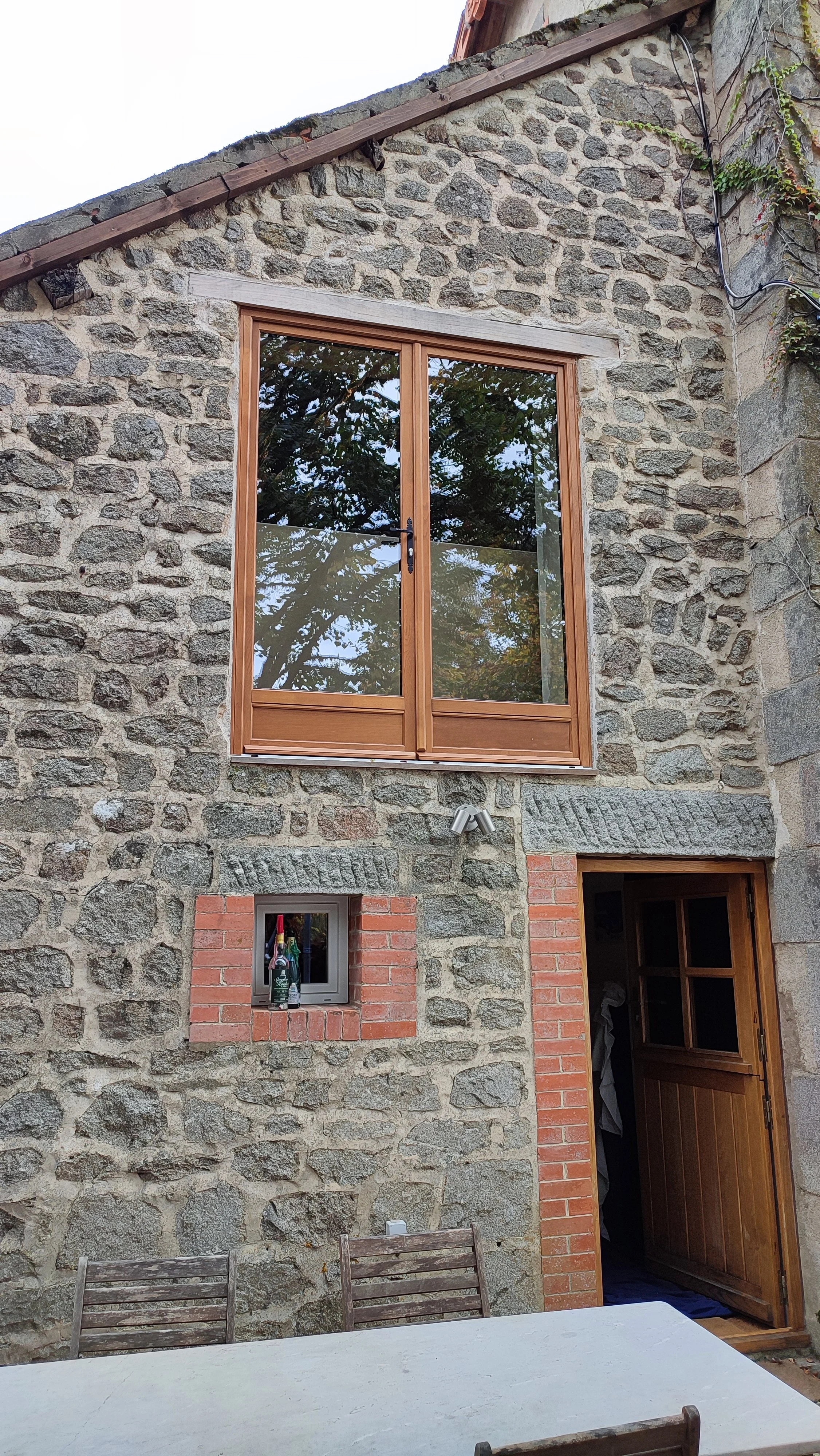 Exterior of a stone house with a wooden framed window on the second floor, a small brick-framed window with bottles, and a wooden door on the ground floor, outdoor dining table with chairs.