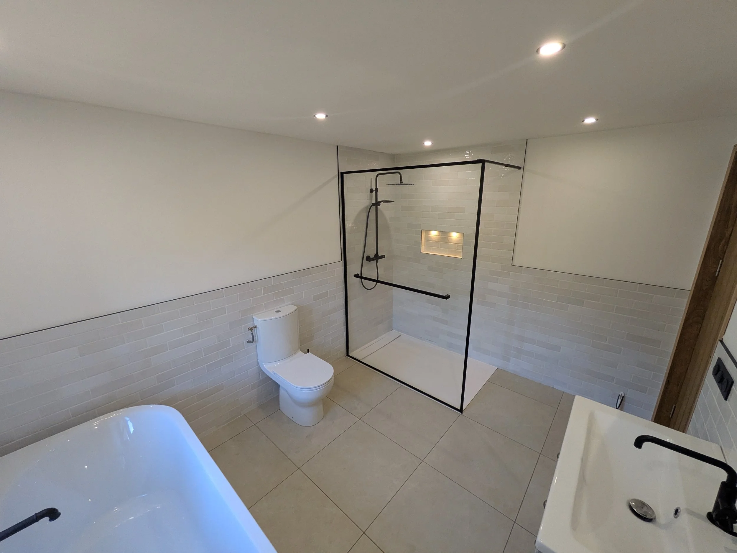 A modern bathroom featuring a white toilet, a white bathtub, and a glass-enclosed shower with black framing, white tiled walls, and a built-in wall niche with lighting.
