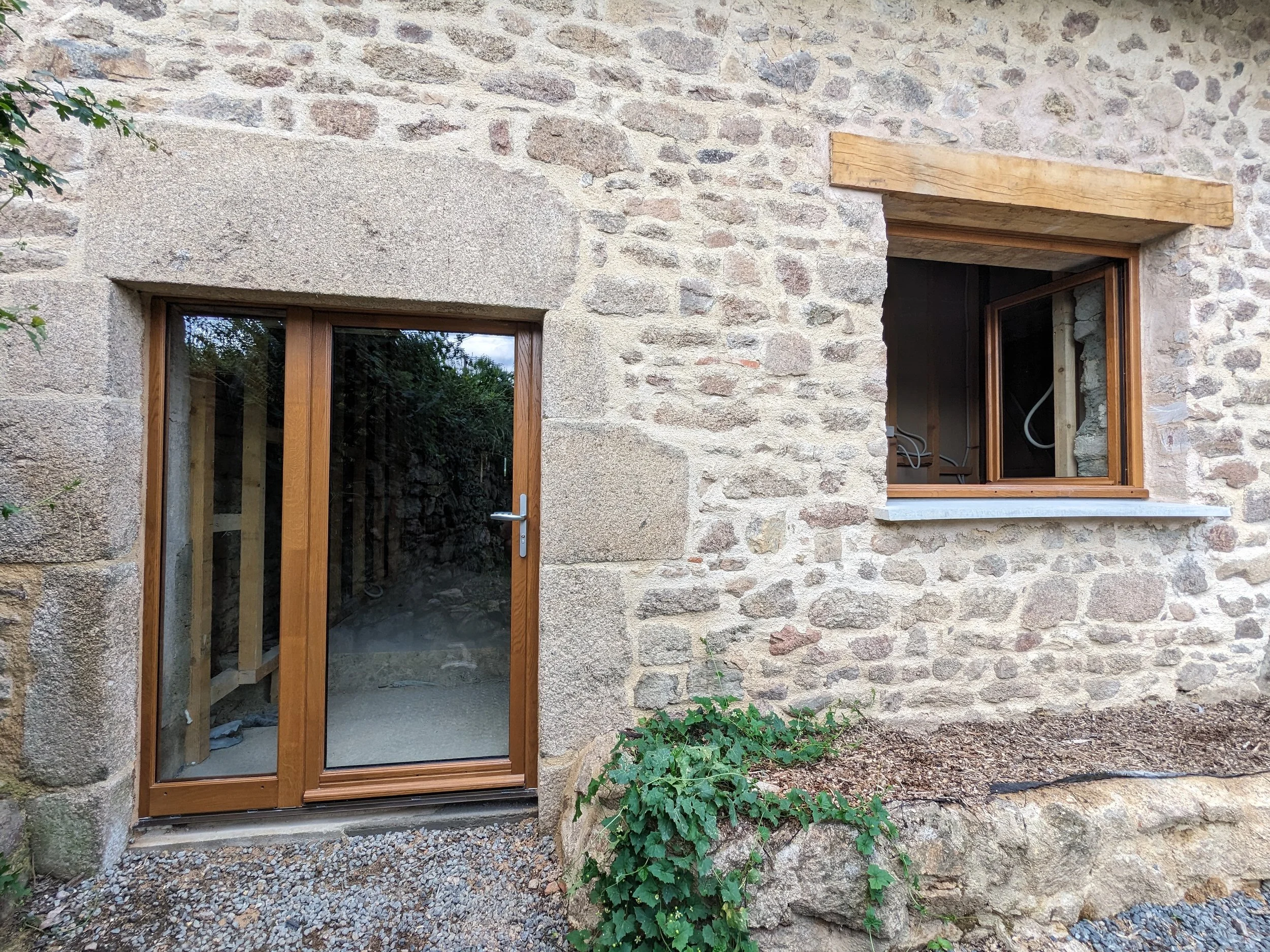 A stone wall exterior of a building with two windows and a wooden lintel above one window. There is a glass door with a wooden frame and handle on the left side. Some green plants and small shrubs grow near the building's base.