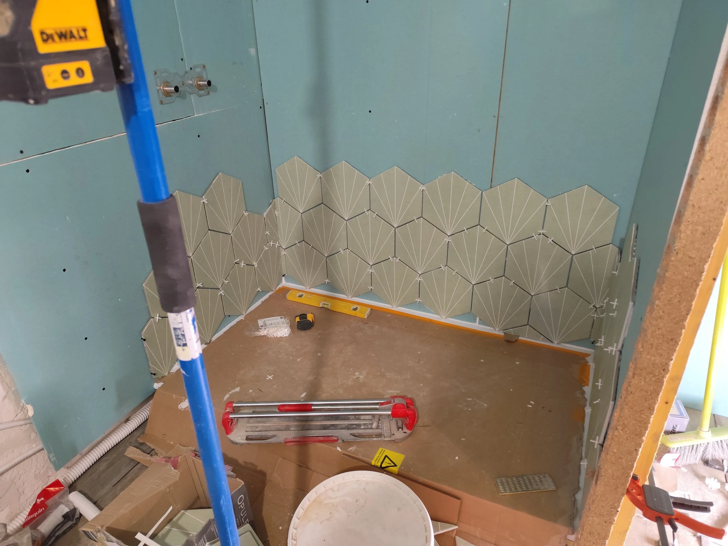 Partial view of a bathroom or utility room under construction, showing green drywall with some hexagonal patterned tiles on the lower part of the wall, construction tools and materials on the floor, and plumbing fixtures.