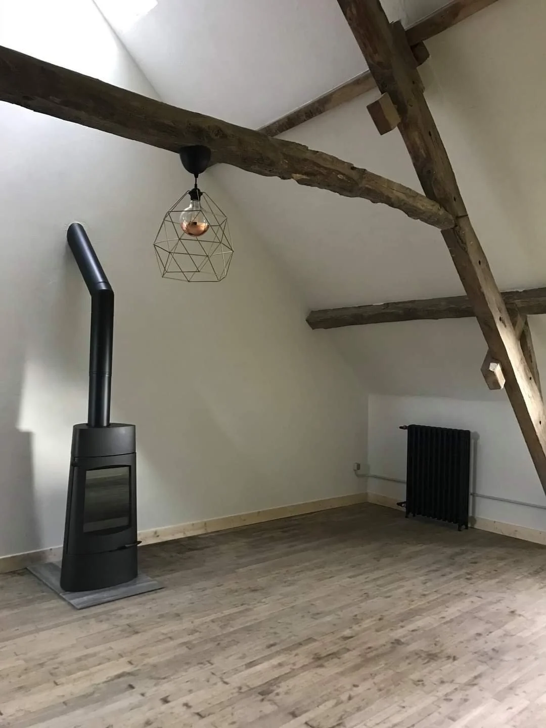 Empty room with wooden beams on ceiling, black stove in corner, radiator on wall, wood flooring, minimalist decor, pendant light hanging from ceiling.