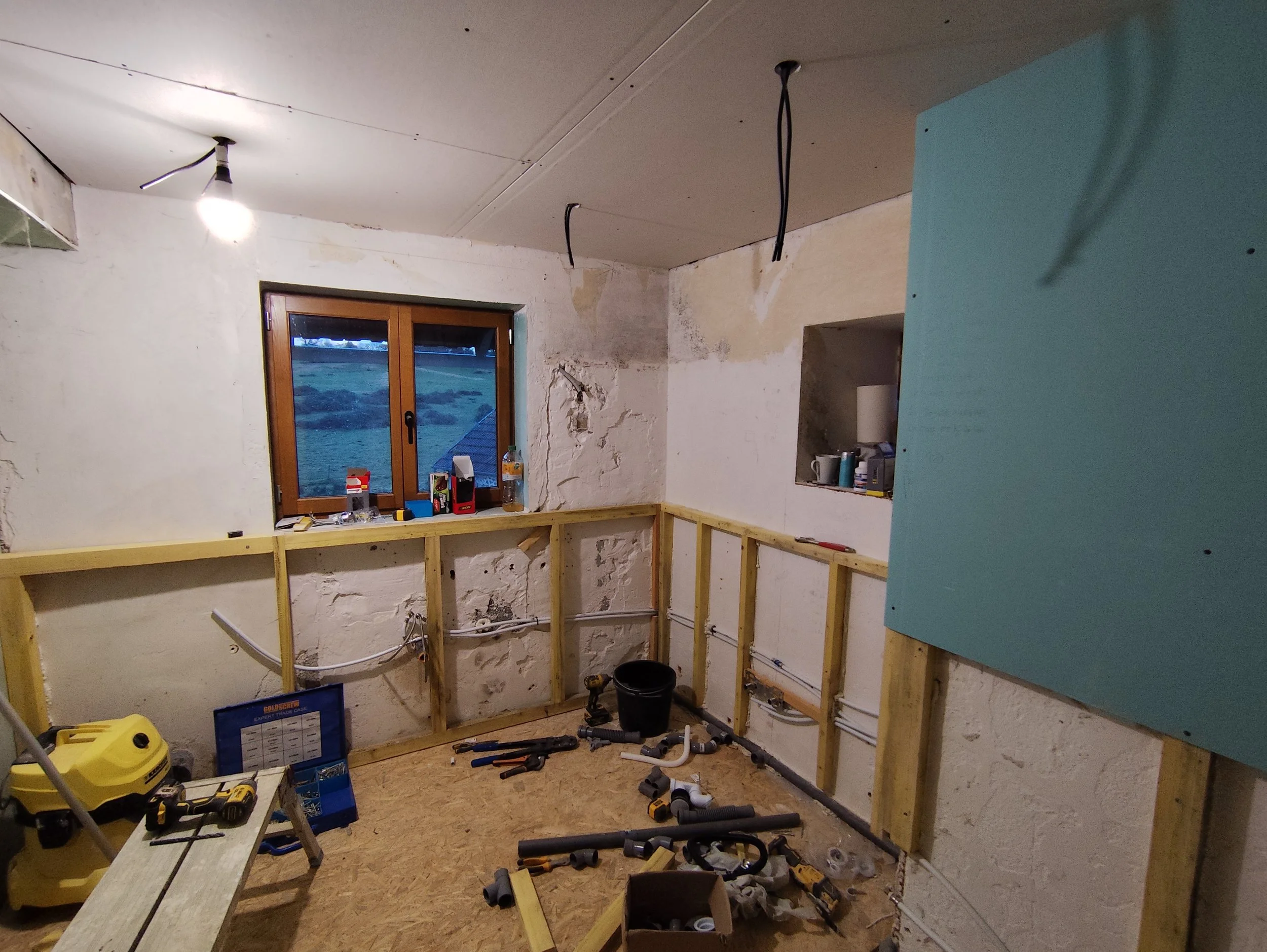 Interior of a room under construction with wooden framing, tools, and pipes on the floor, and a window showing an outdoor landscape.