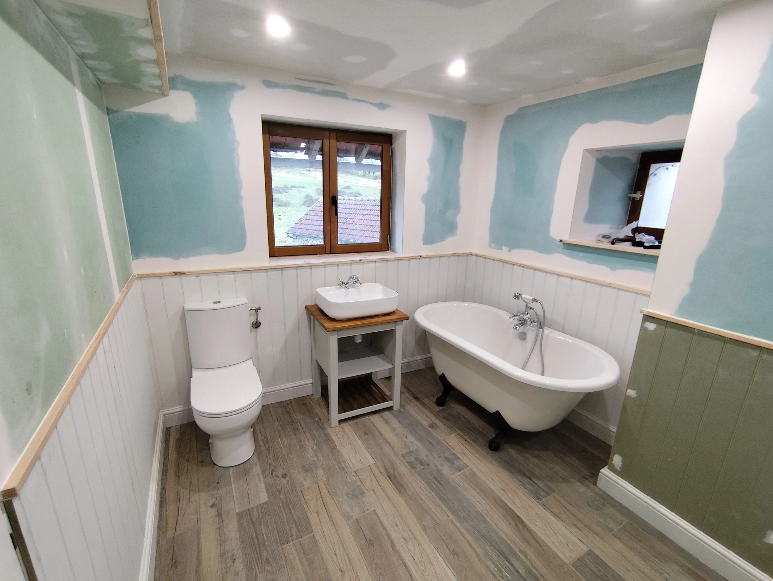 Bathroom with green walls, white wainscoting, a bathtub, a toilet, and a sink with a small wooden stand. There are two windows, one larger and one smaller. The ceiling has recessed lighting.
