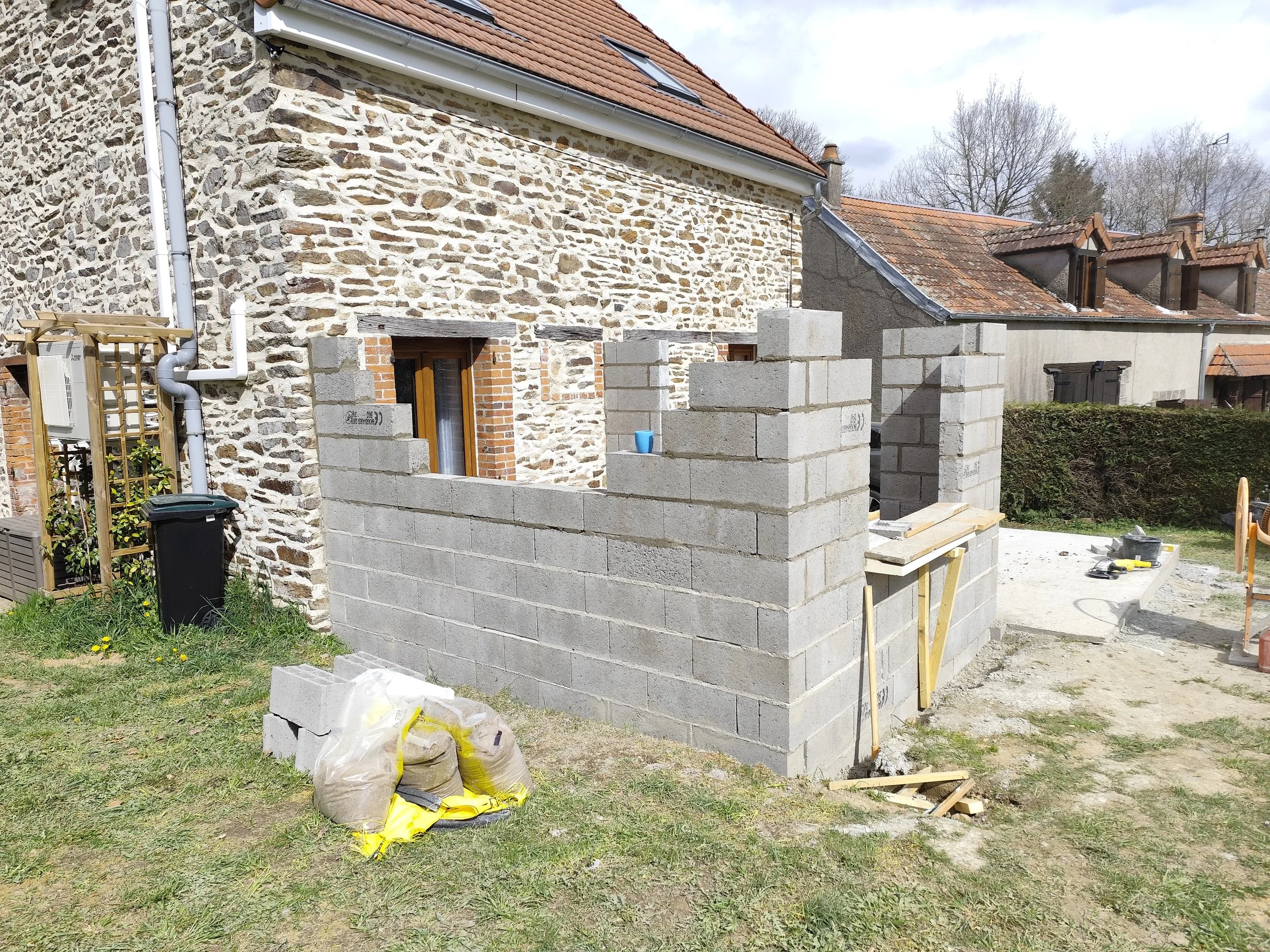 A house with a stone wall exterior undergoing construction, with a new cinder block wall being built in the foreground, construction tools and bags of materials are visible on the grass.