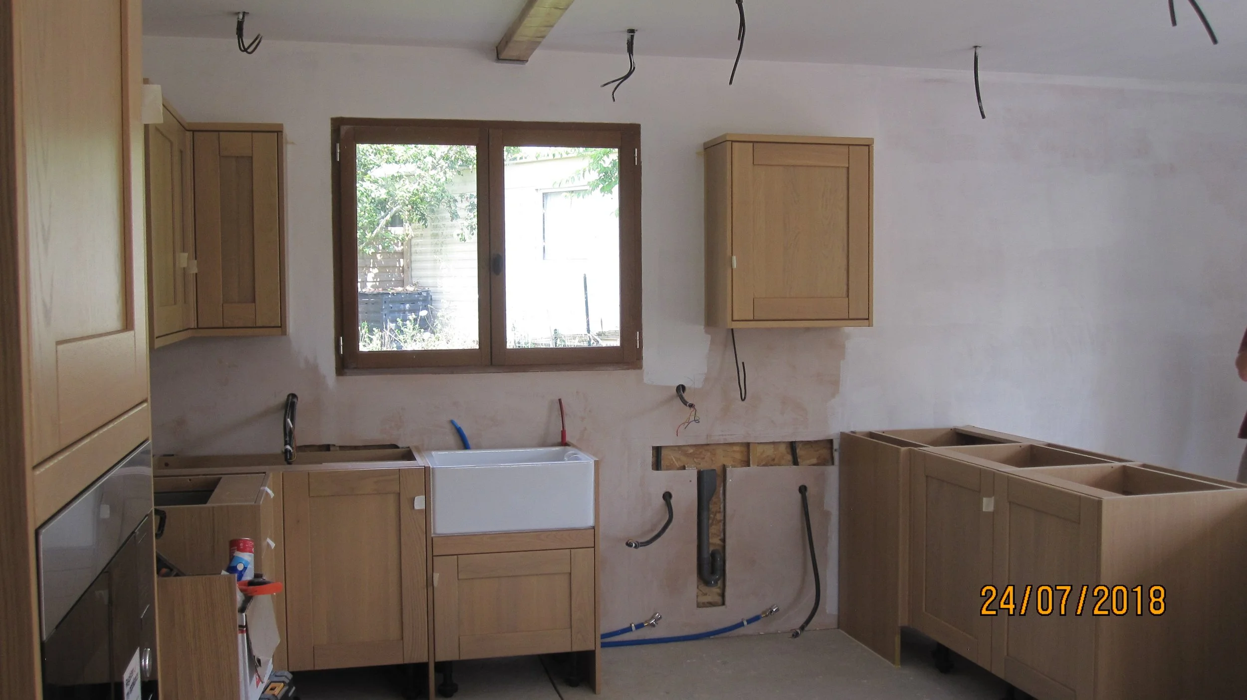 Kitchen under renovation with wooden cabinets and no appliances installed yet. Electrical wires hang from the ceiling, and plumbing is exposed under the sink area. A window provides natural light.