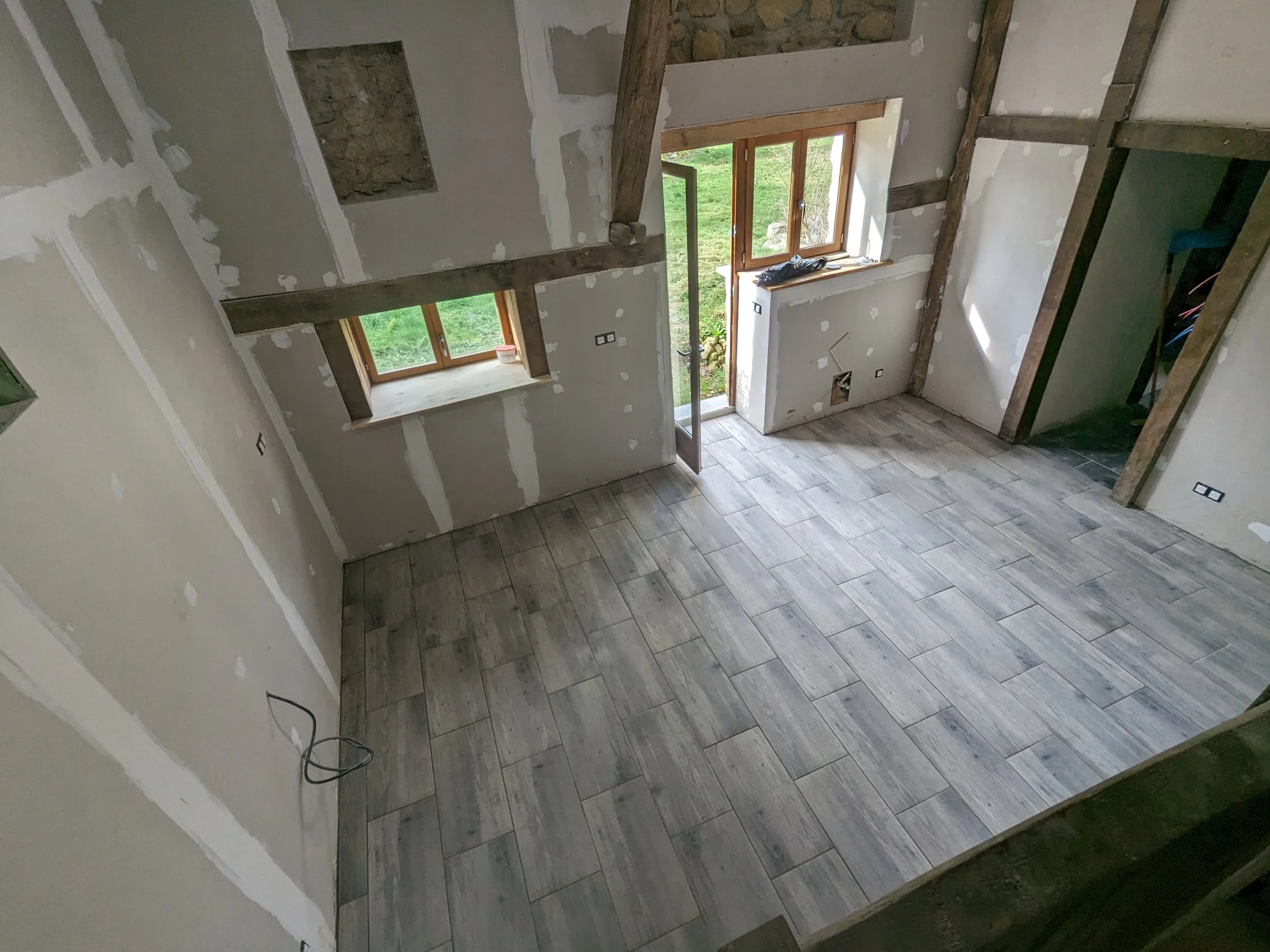 An interior room under construction with unfinished drywall walls, visible wooden beams, a window with a view of green grass outside, and tiled flooring. There are also some electrical wires and outlets visible.