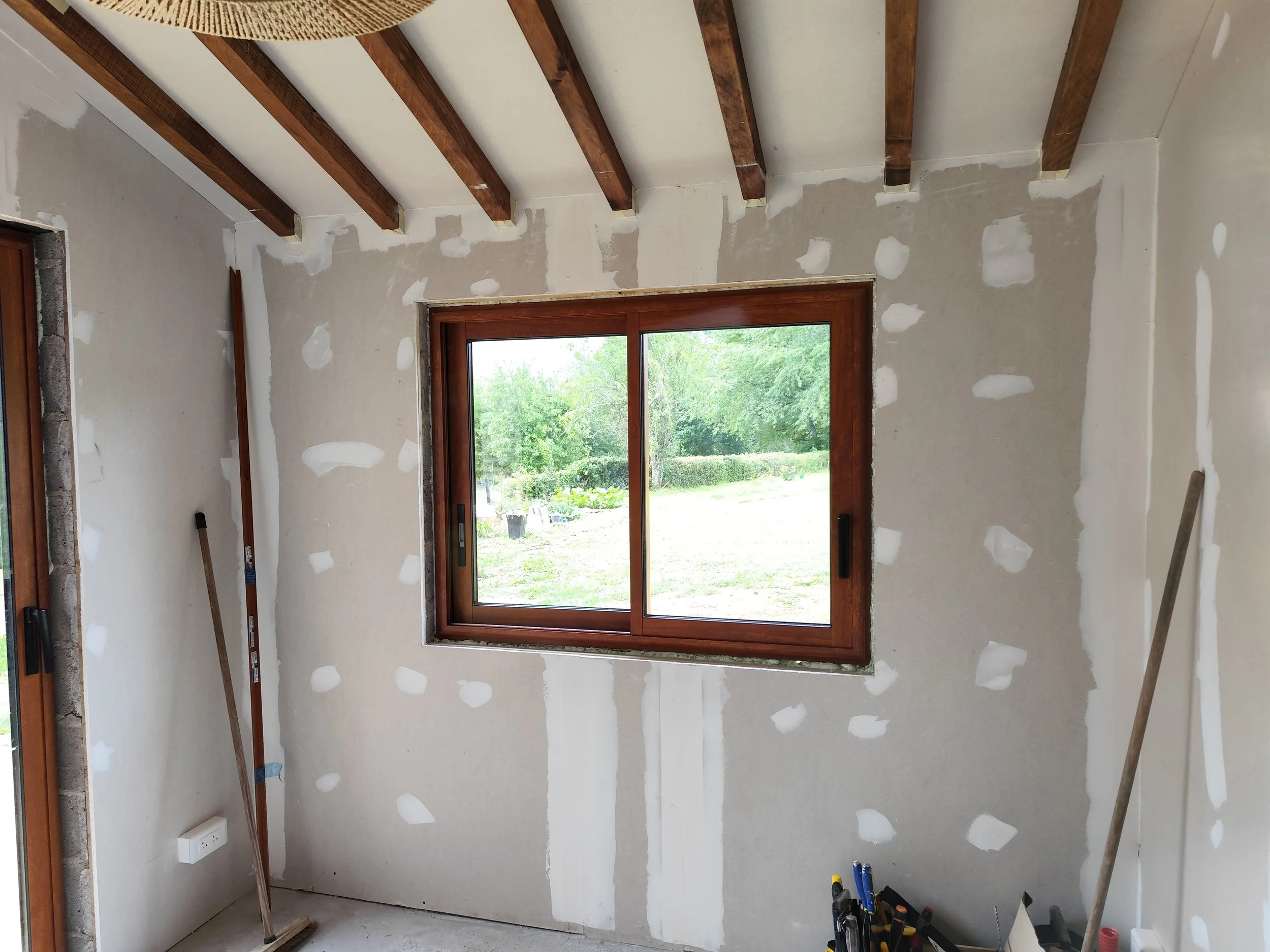 Interior of a room under renovation with unfinished drywall walls, wooden ceiling beams, and a large window showing greenery outside.
