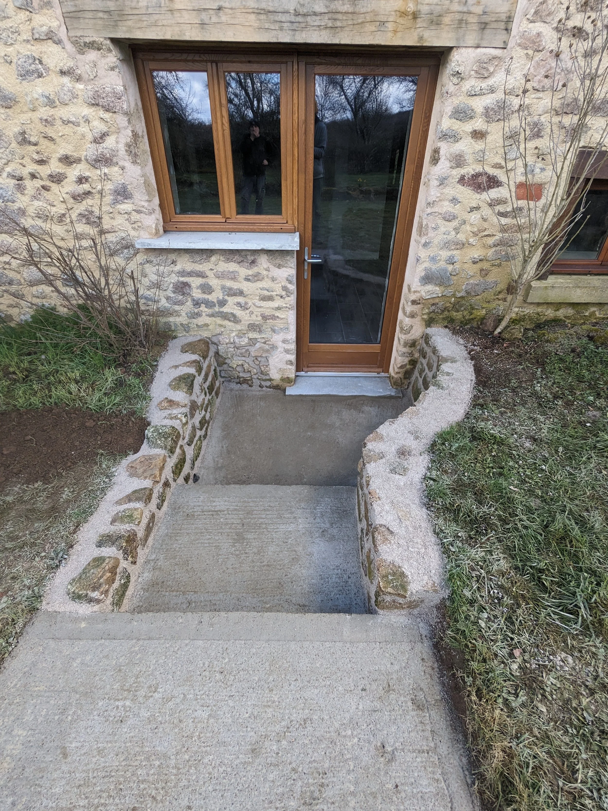 Exterior stone building with entrance door and stairs, leading down to a concrete sidewalk, bordered by dirt and grass with a small tree on the right.