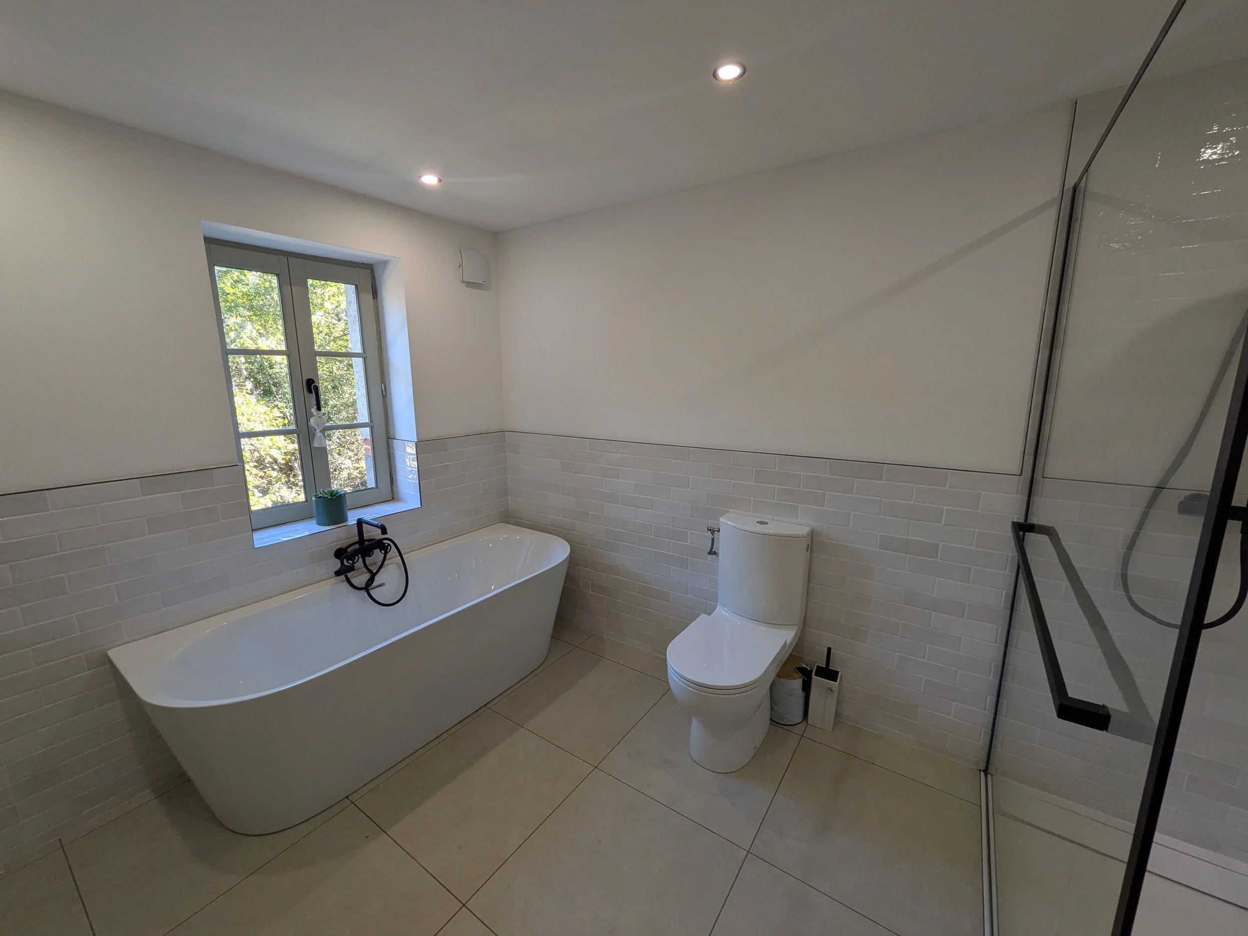 A modern bathroom with a white bathtub under a window, a toilet, a small potted plant on the window sill, and a shower area with a glass door and black fixtures.