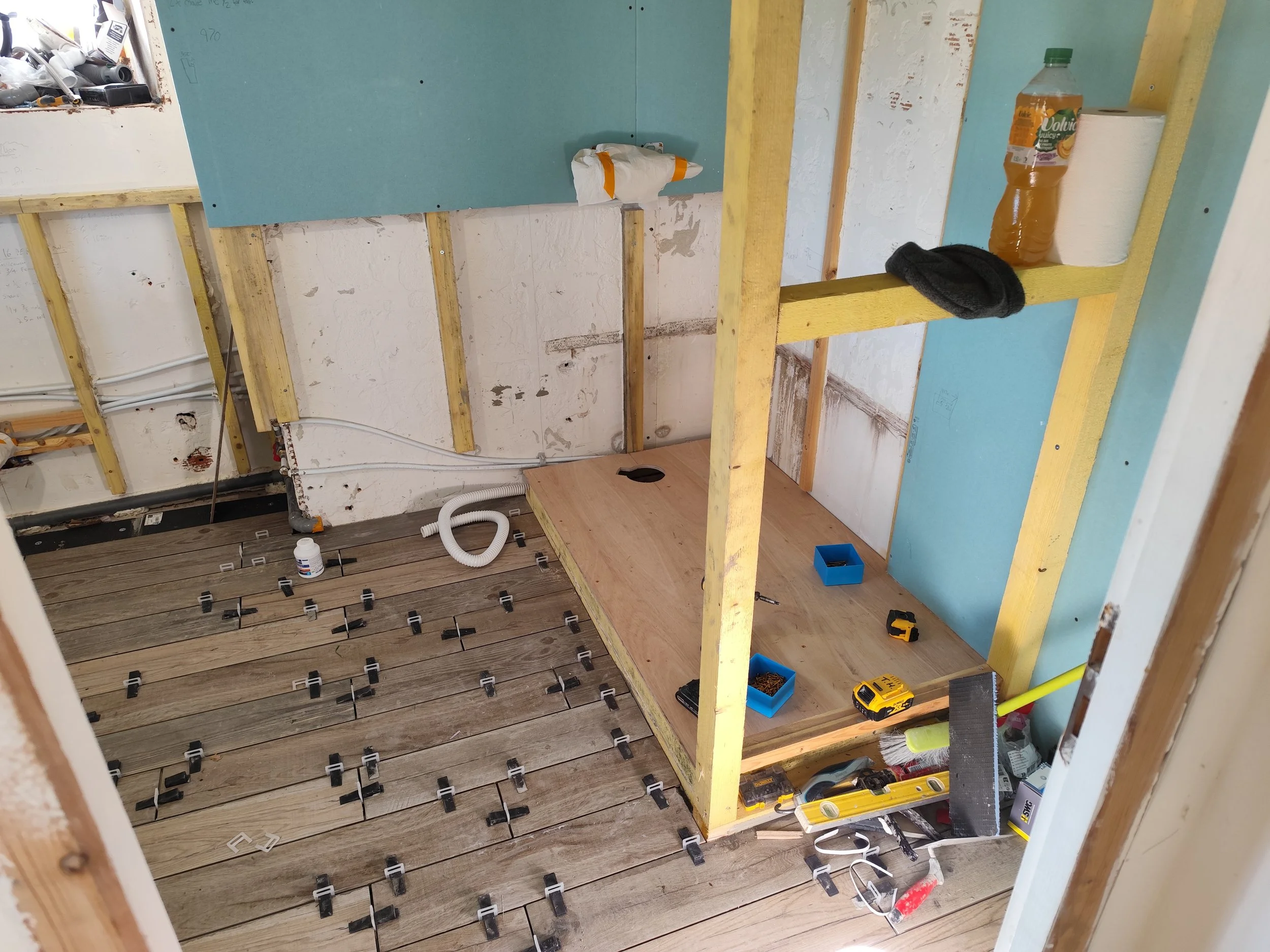 Interior of a room under construction with exposed framing, partially installed flooring, and tools scattered around. An unfinished wall with insulation and wiring is visible, along with construction materials and equipment.