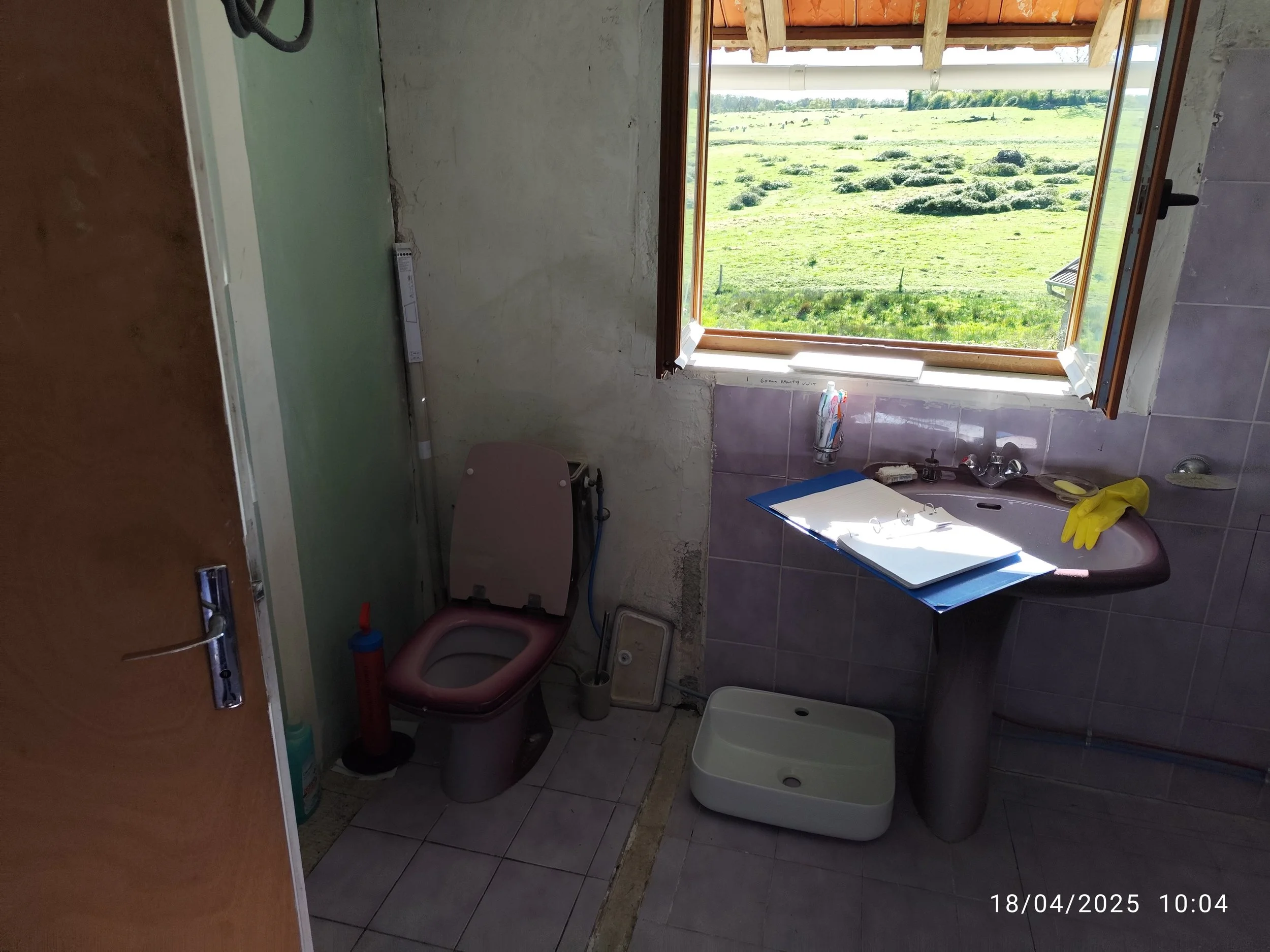A bathroom with a window overlooking a grassy field with hills. The bathroom has a pink toilet, a sink with a mirror above it, and a sink basin on the floor. There is a yellow glove on the sink. The walls are partially tiled and mostly unfinished, wi