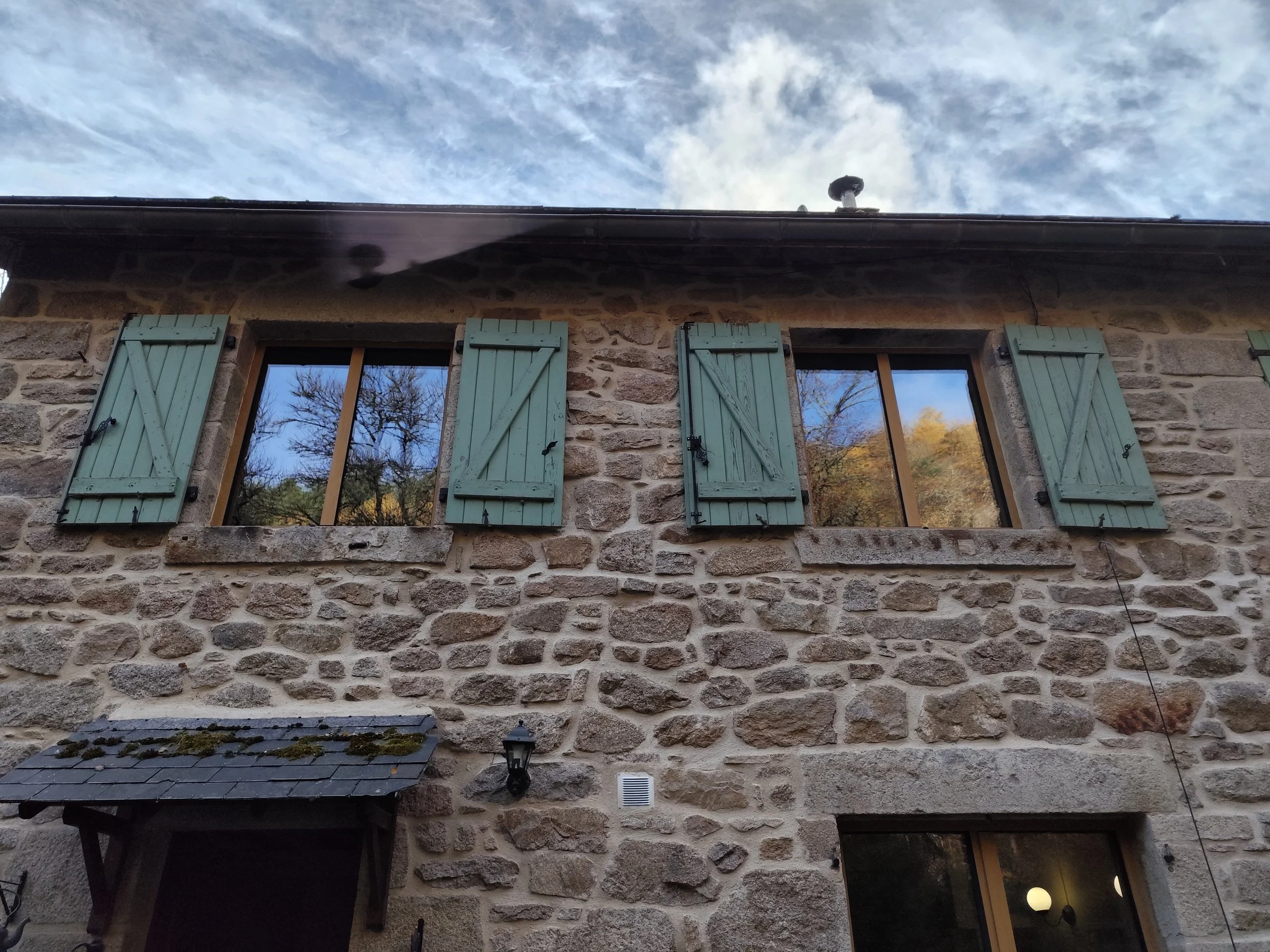 A stone house with three windows with closed green shutters, a black roof with moss, a small black outdoor table, a lantern, a white vent, and a partial view of a tree and sky in the reflections.