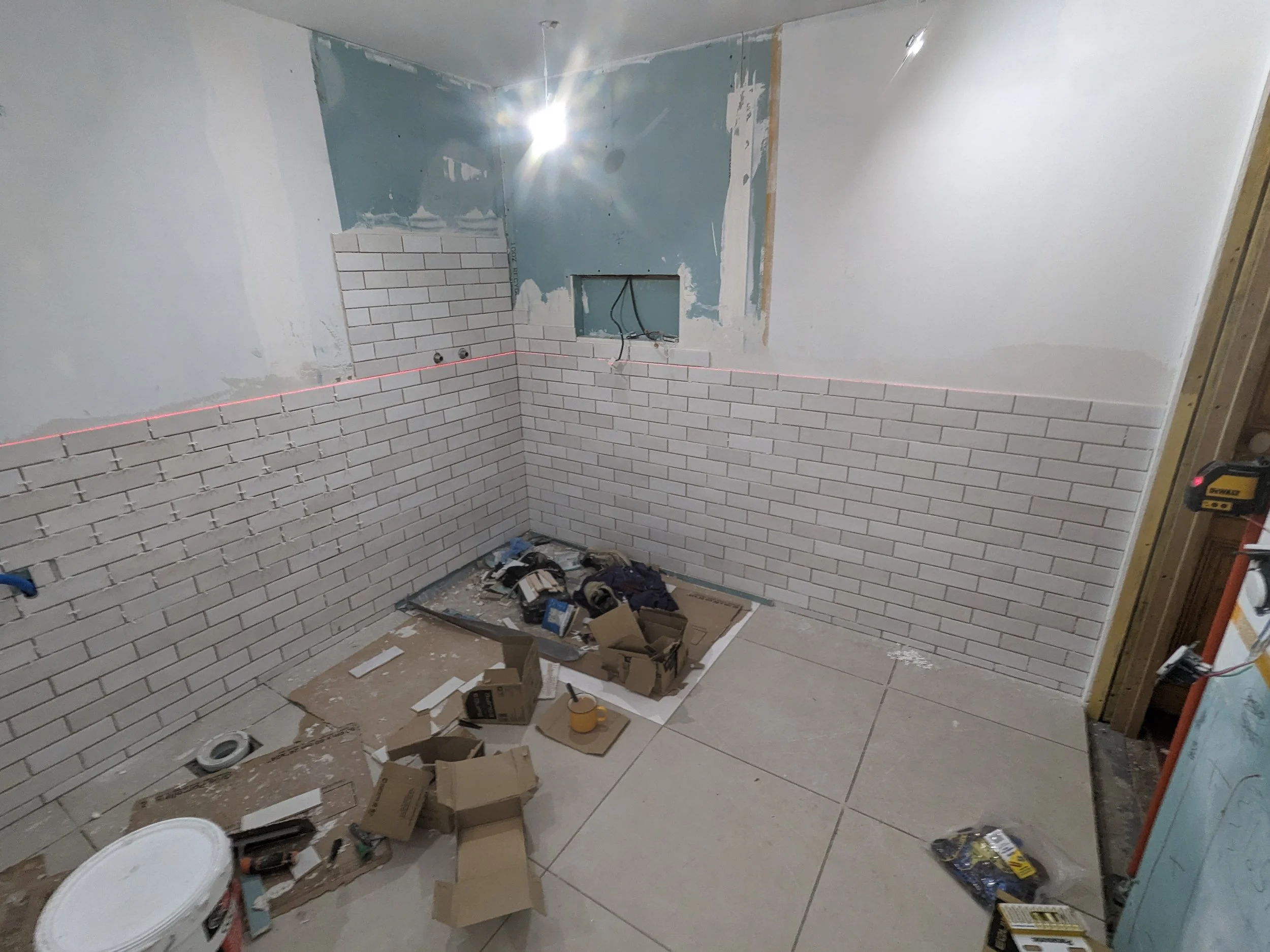 Room under renovation with white brick wall tiles partially installed, construction tools, boxes, and supplies scattered on the floor, and a laser level line on the wall.