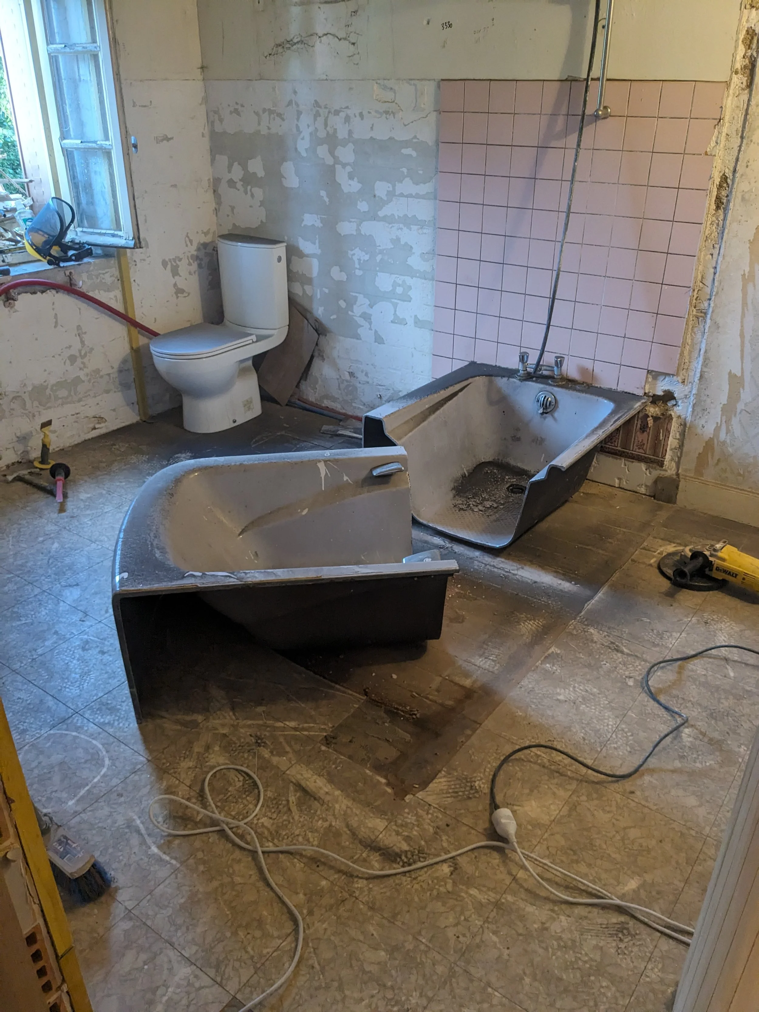 A bathroom under renovation with a toilet, two bathtubs, construction tools, and exposed walls and flooring.