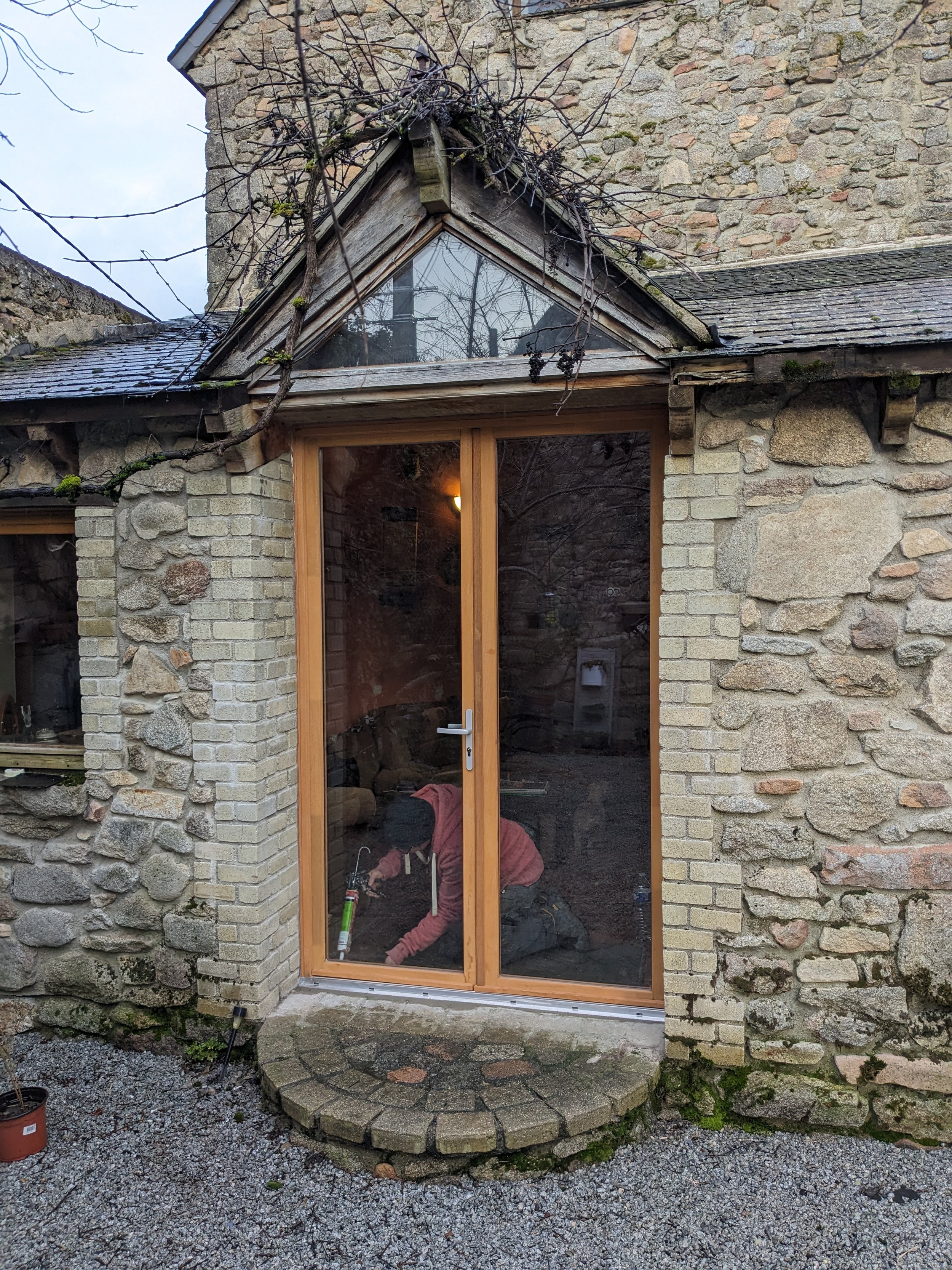 Worker installing a large glass door in the stone house's brick wall with a stone step and gravel ground in front.