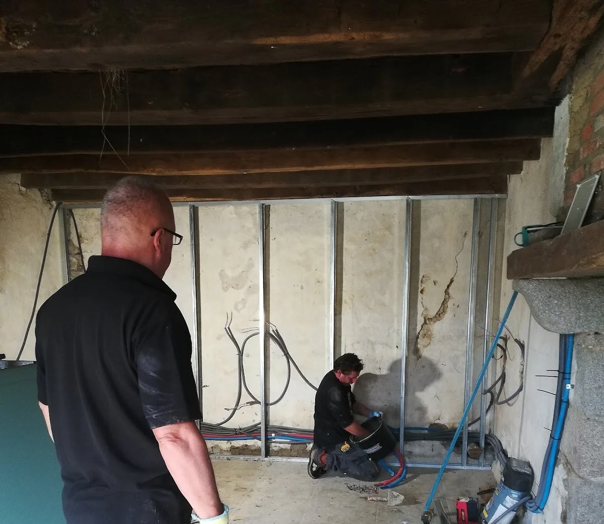Two men working on electrical wiring in a room with exposed wall studs and plumbing.