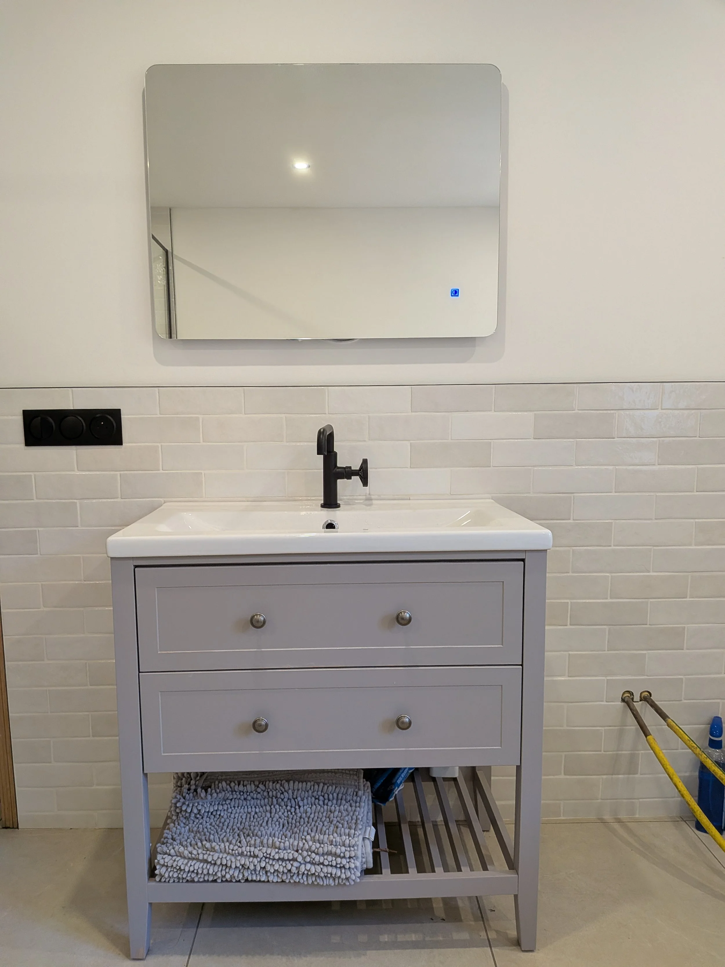 A small bathroom vanity with a black faucet, a white cabinet with two drawers, and a shelf below holding a textured beige towel and various items. Above it is a wall-mounted rectangular mirror with rounded edges, reflecting a ceiling light and part o