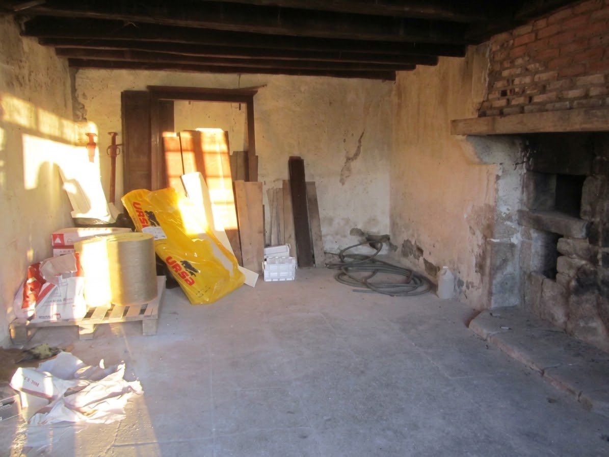 An unfinished room with construction materials, including insulation, wood, and bricks. The room has a brick and plaster fireplace and sunlight streaming through a window.
