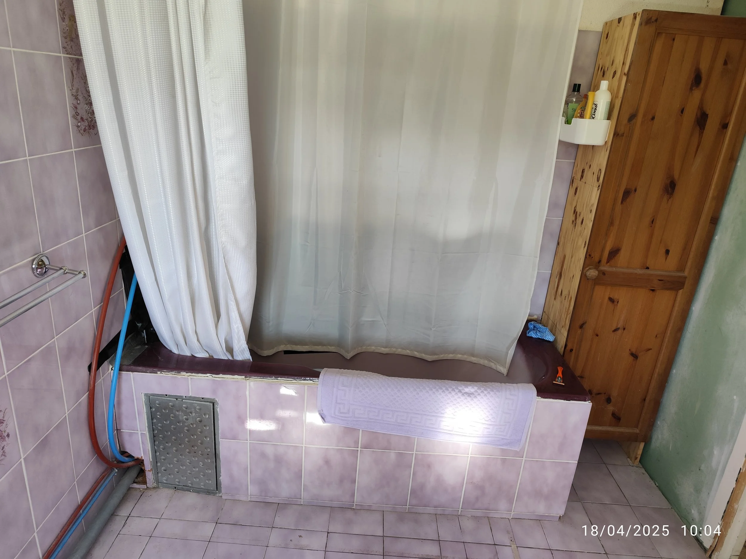 A bathroom with pink tiled walls and floor, a white shower curtain, a wooden cabinet, a towel hanging, and plumbing pipes visible. The photo was taken on April 18, 2025, at 10:04.