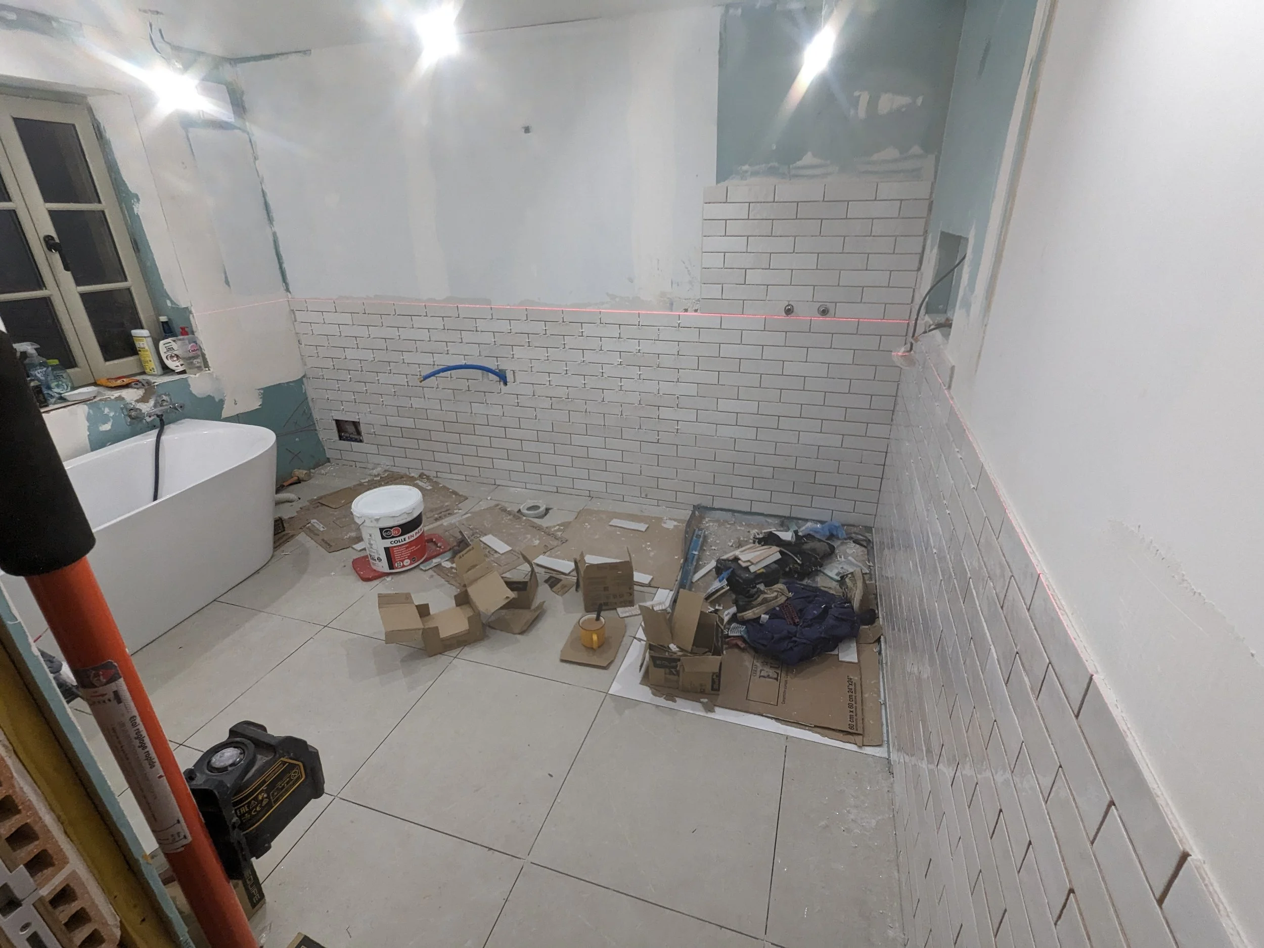 A room under renovation with partially painted walls, stacks of cardboard boxes, construction tools, and supplies scattered on the floor. Red laser level line running across the wall, unfinished electrical outlets, and a bathtub visible.