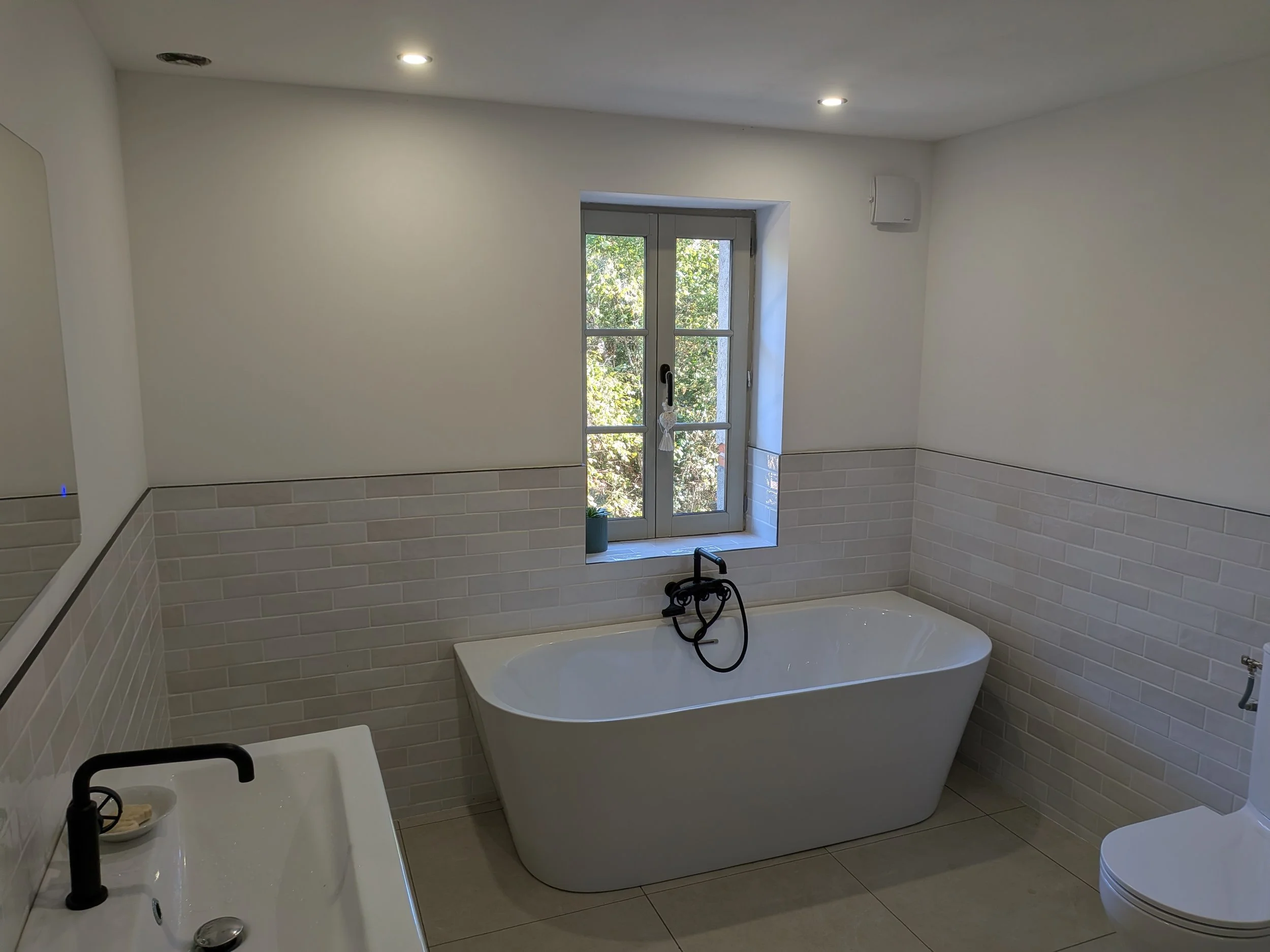 A bathroom with white walls, a large window with a view of trees, a bathtub with a black faucet, and a small white sink with a black faucet.