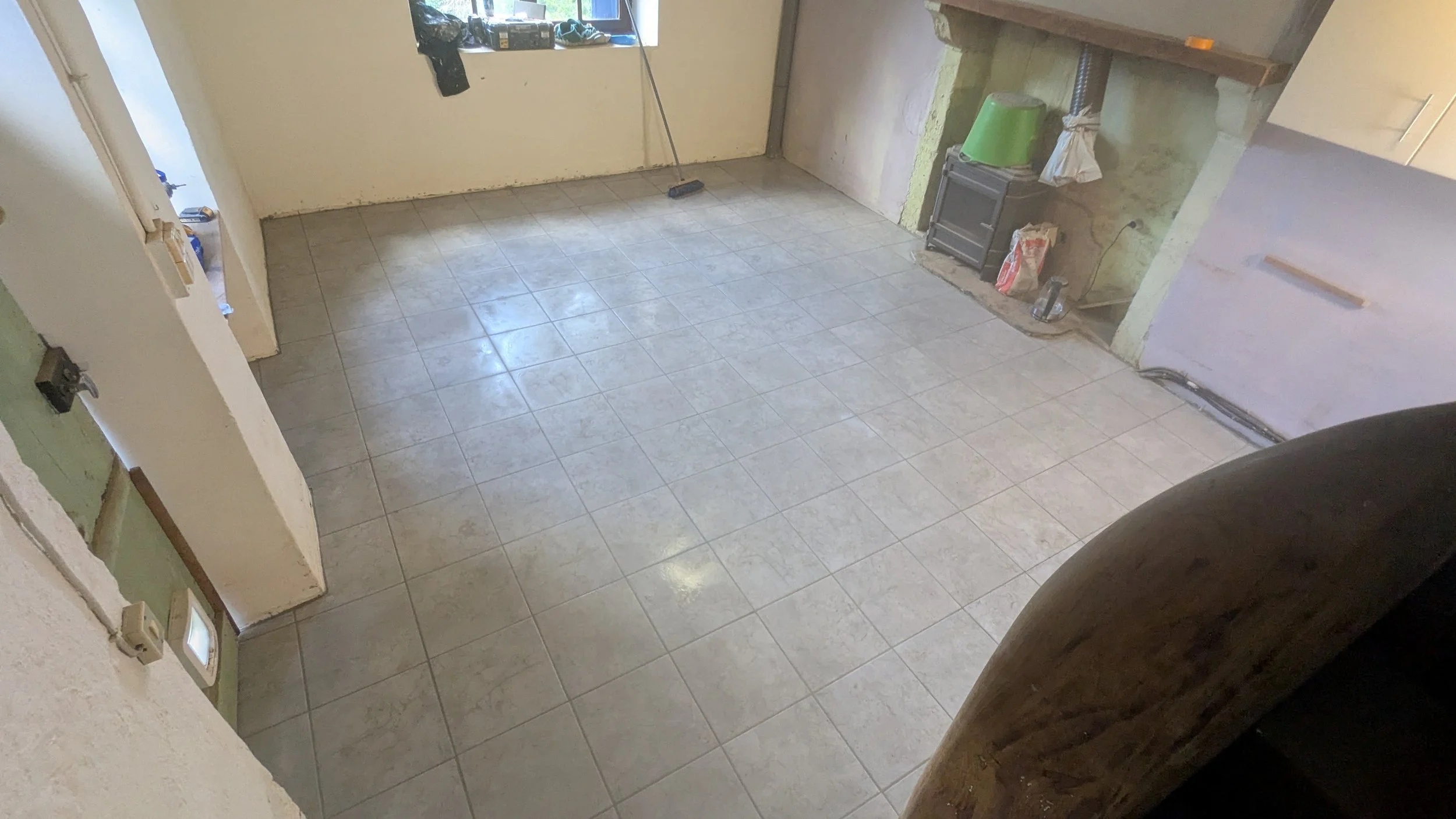 Empty room with tiled floor, unfinished walls, a window with items on the sill, a broom leaning against the wall, and construction supplies near an open wall area.