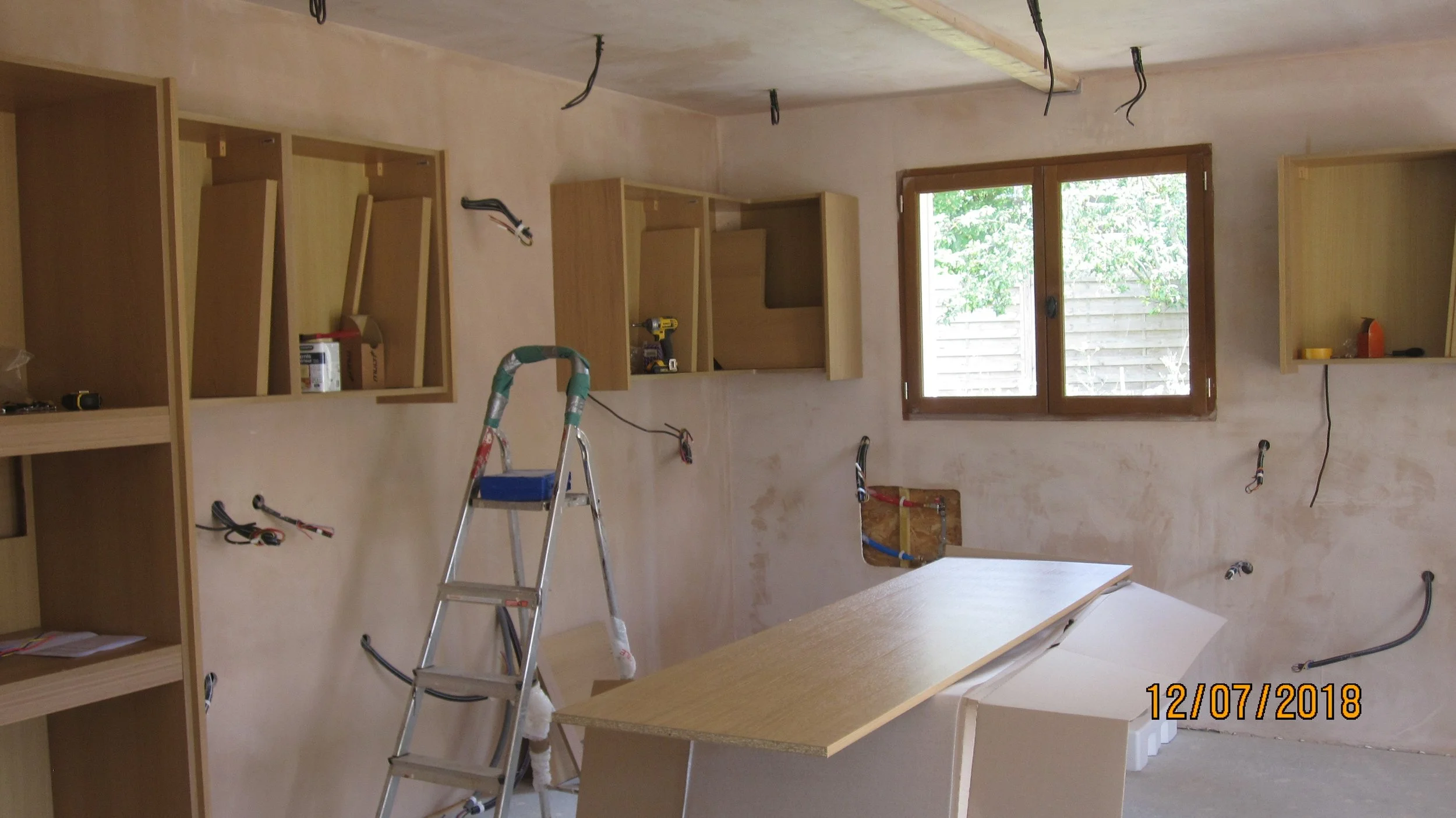 Interior of a room under construction with unfinished walls, exposed wiring, and wooden shelving units. There is a step ladder in the center and a window showing greenery outside.
