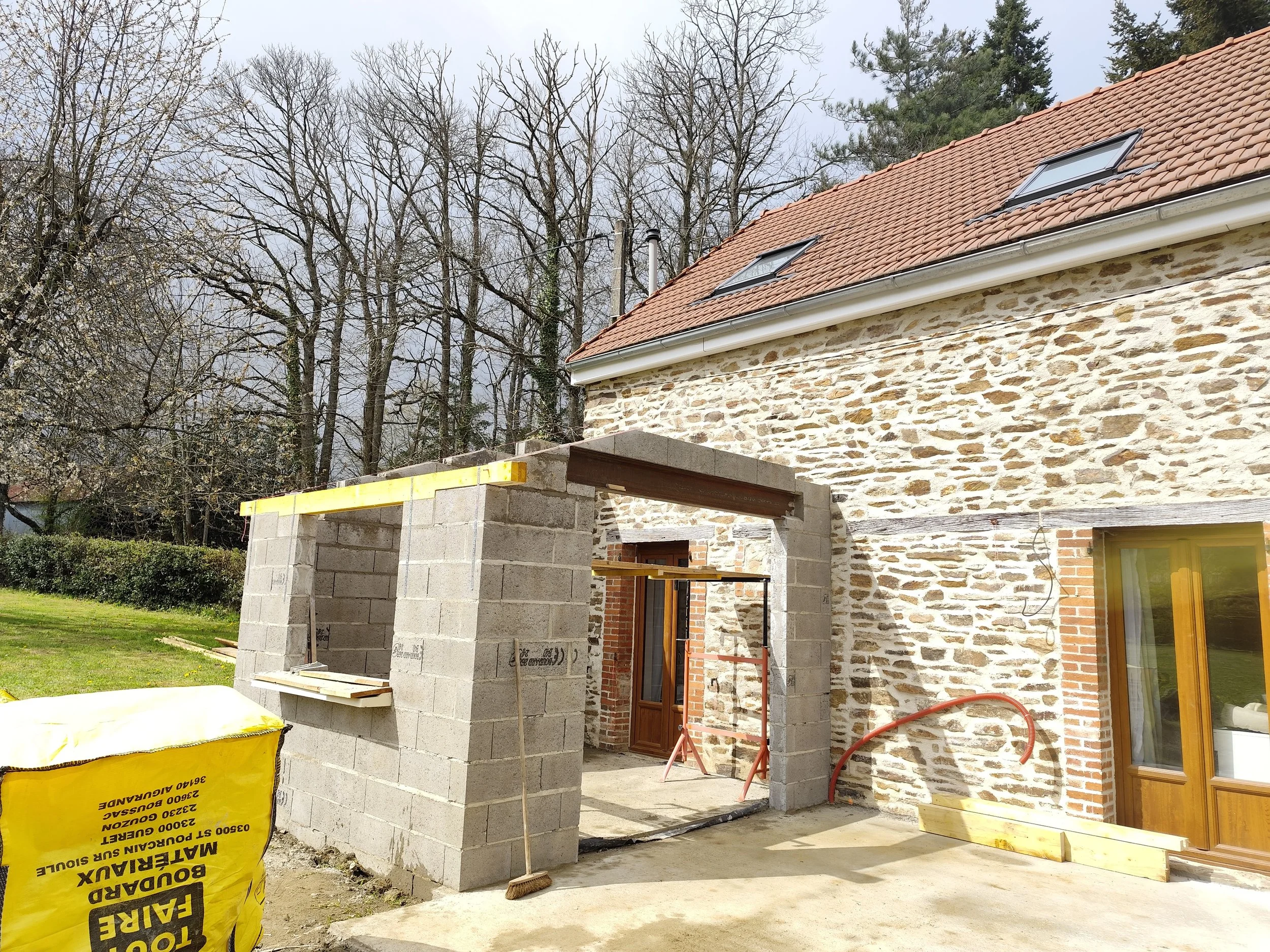 A house under construction with a brick and stone exterior. There is a concrete block structure in progress, and yellow bags of construction materials nearby. The house has two skylights on the roof, and the yard has grass and trees.