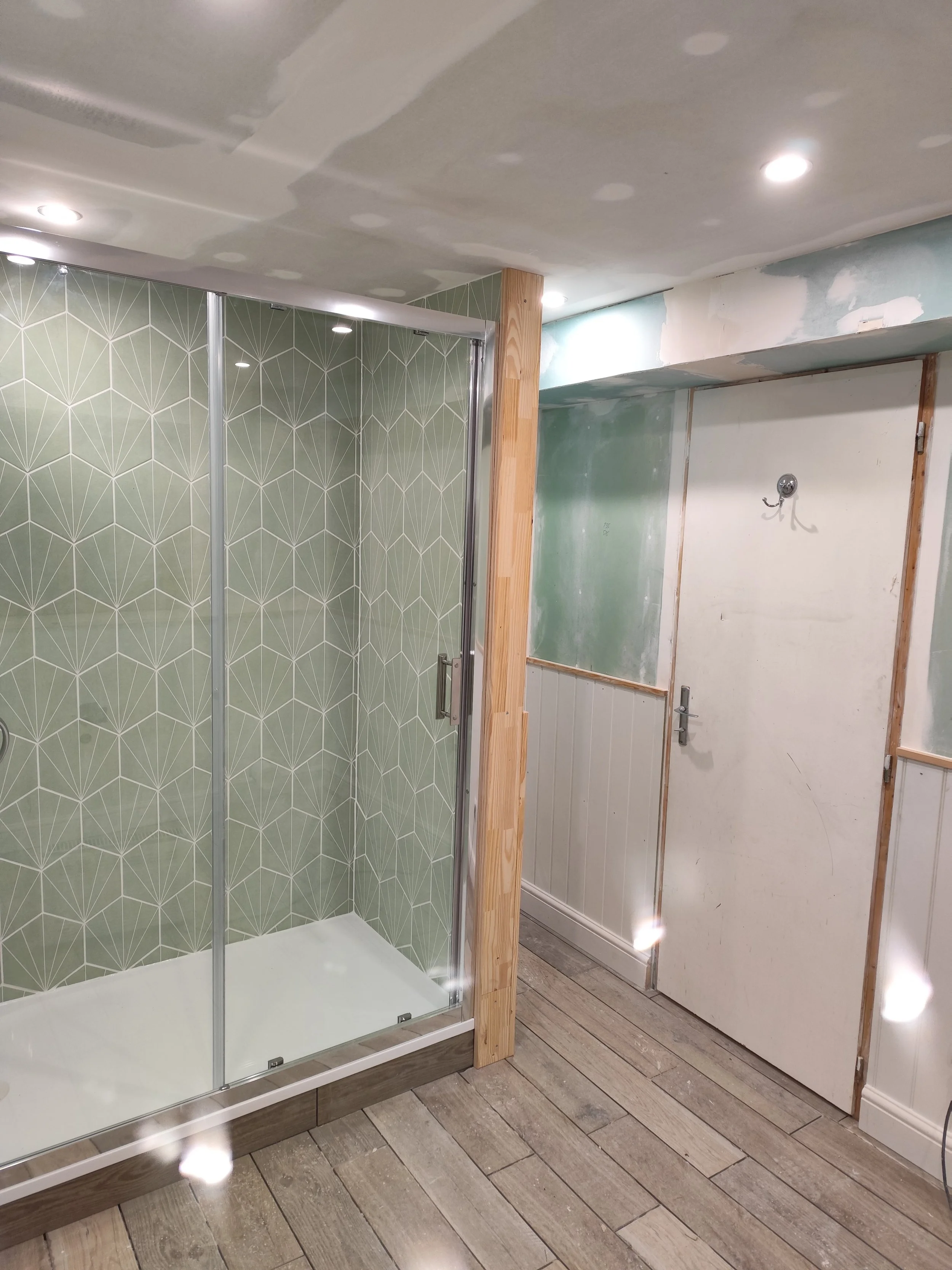 Interior of a bathroom under construction featuring a corner shower with patterned tiles, unfinished walls, a temporary door, and wooden framing, with wooden flooring.