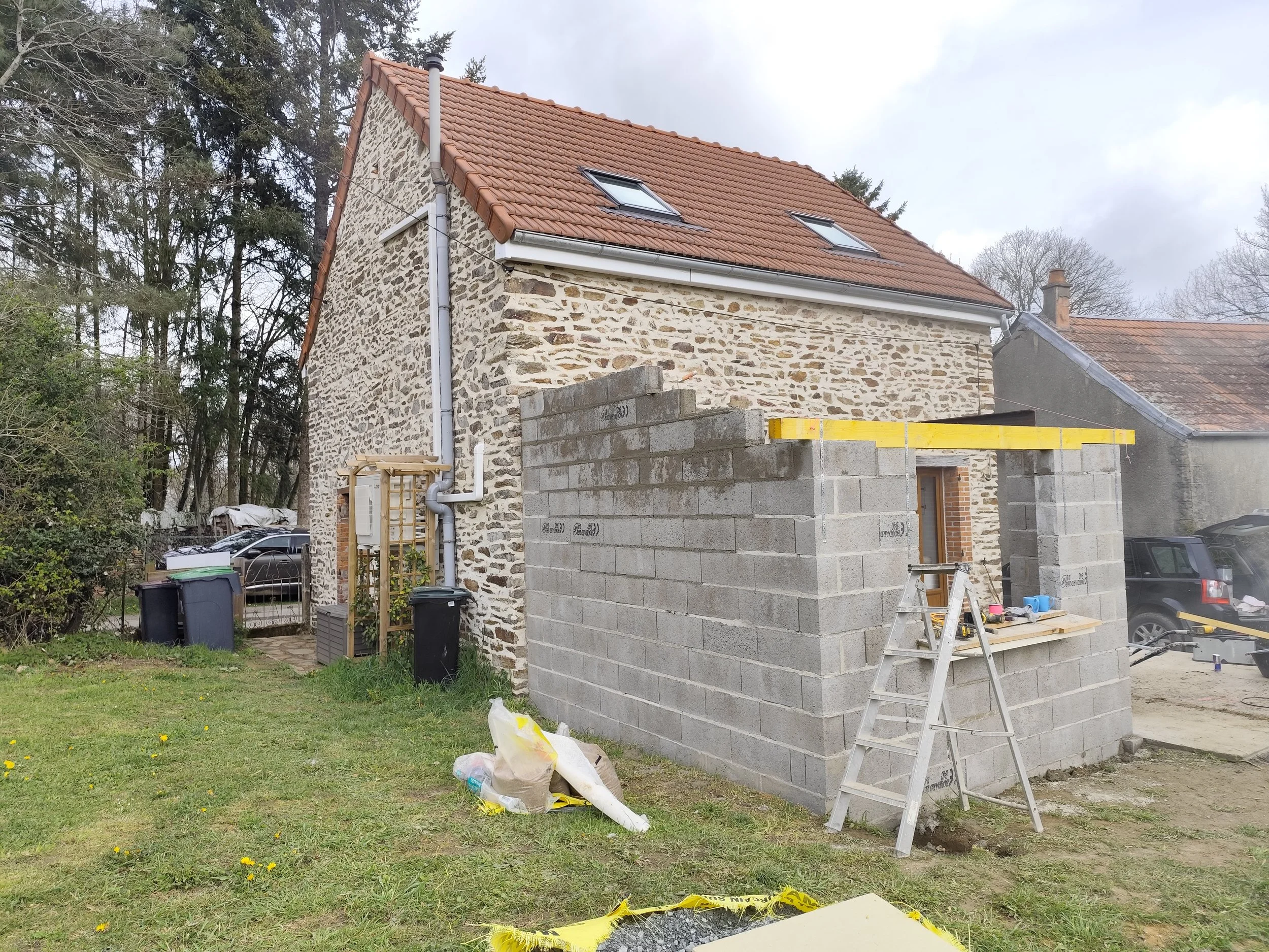 A house under construction with stone walls and a red tiled roof. Construction materials and tools are visible, including cinder blocks, a ladder, and bags of construction supplies. There are trees in the background and a grassy lawn in the foregroun