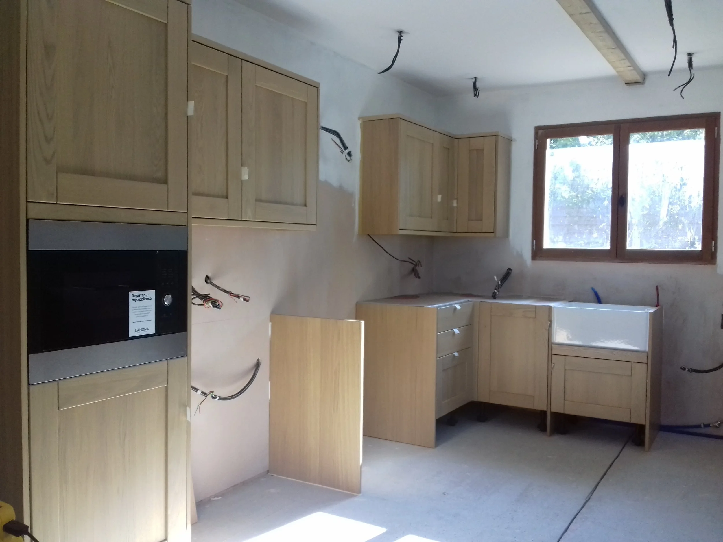 Interior of a kitchen under construction with wooden cabinets, a window, and exposed wires hanging from the ceiling.