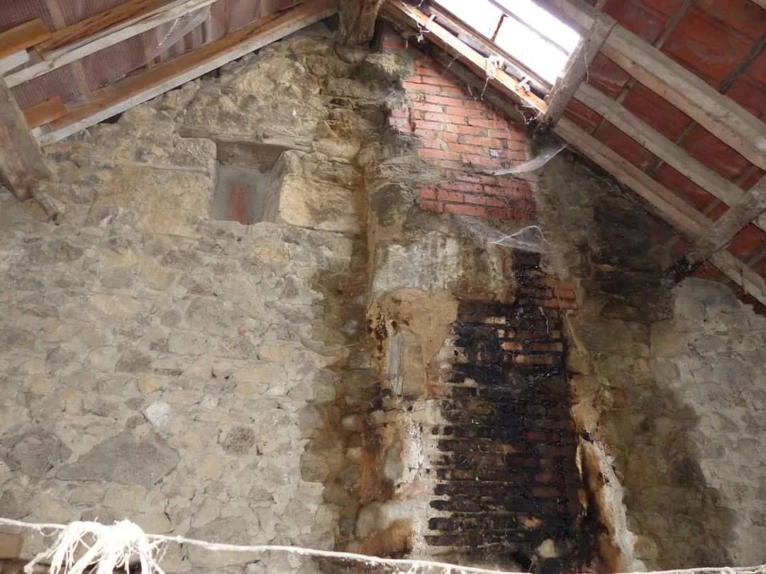 Inside a stone and brick chimney with some soot and cobwebs, located in an old building. The roof has exposed wooden beams and some sunlight coming through a small window or vent at the top.