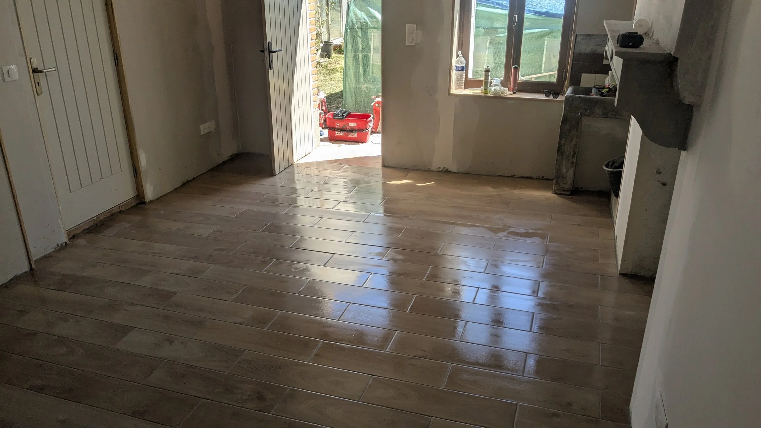 A room with newly installed wood-look tile flooring, an open door leading outside, and a window near the door allowing natural light in. Some construction tools and supplies are visible outside and on the windowsill.
