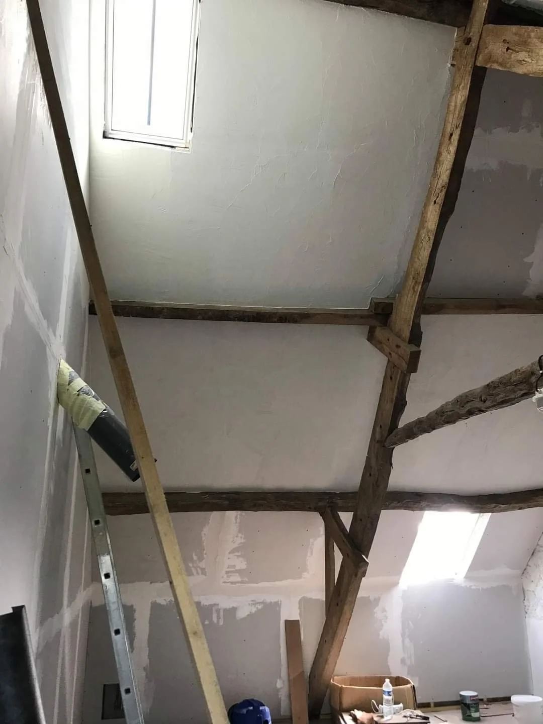 Interior of a room under construction with exposed wooden beams, unfinished walls, a ladder, and construction supplies on the floor.