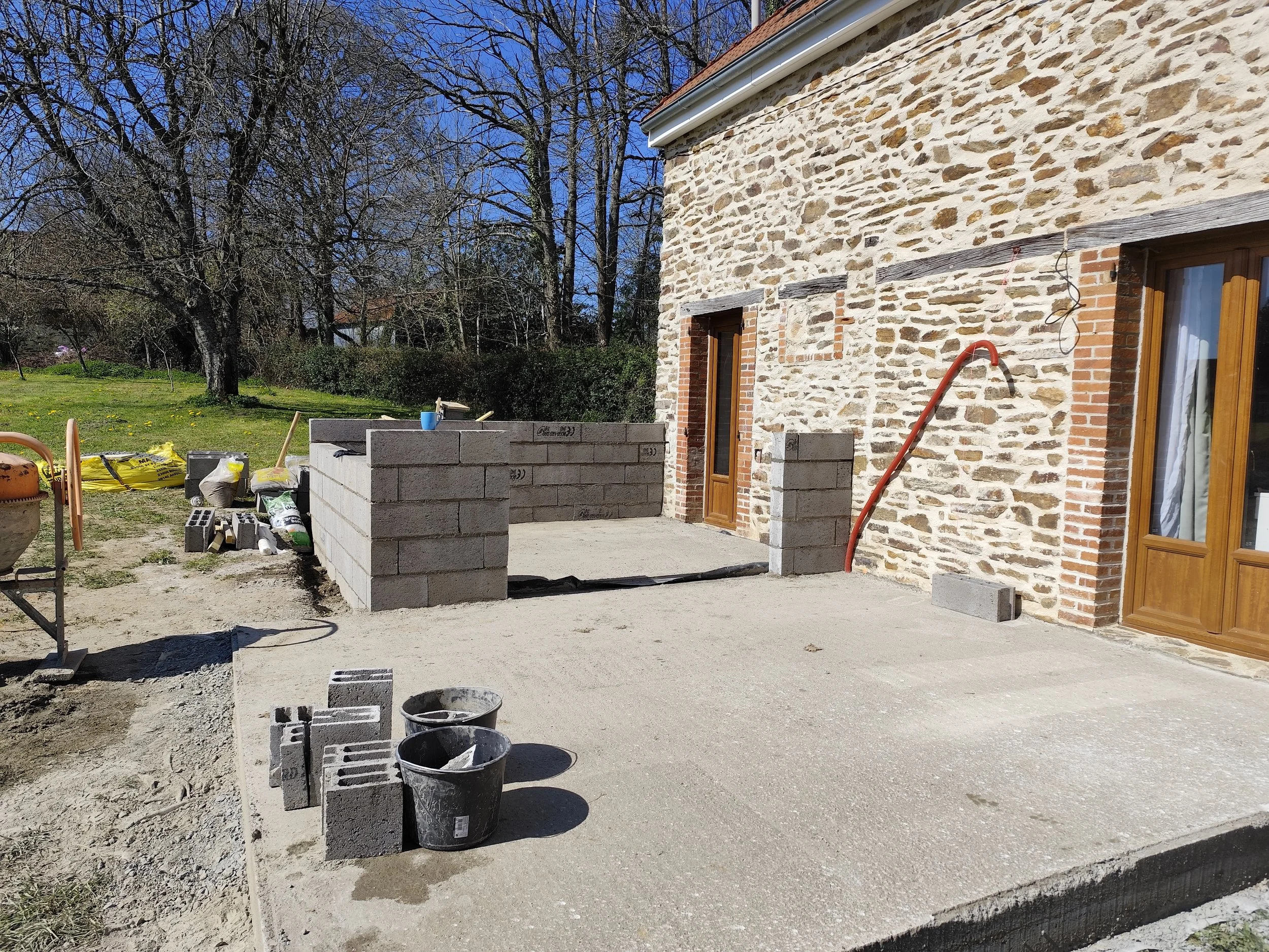 Construction site with a stone wall on the right side, a partly built concrete block wall, construction supplies, trees, and a clear blue sky in the background.