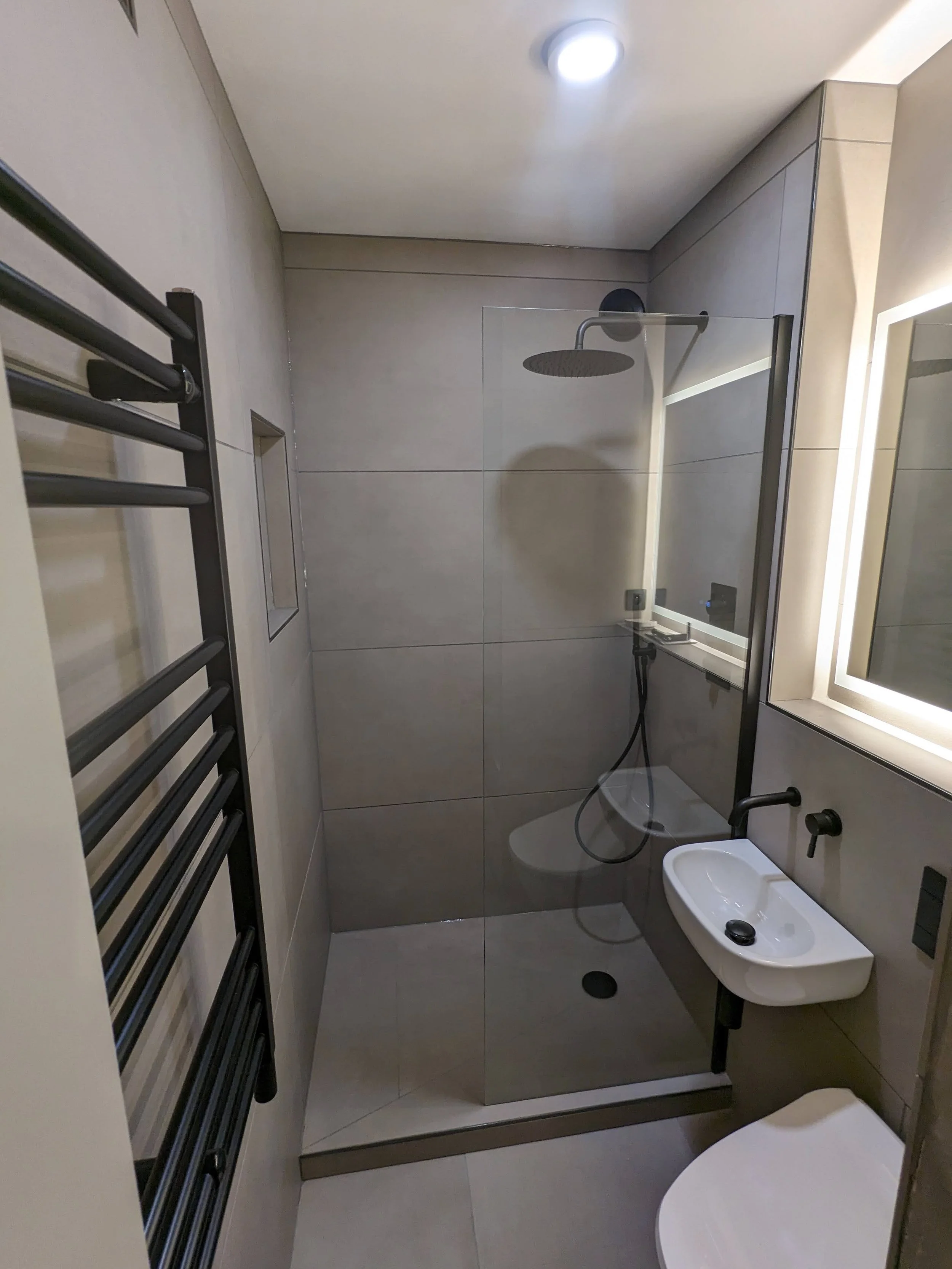 Modern bathroom with gray tiled walls, a walk-in shower with a rain shower head and glass partition, a wall-mounted white sink, illuminated mirror, black towel rail, and a toilet.