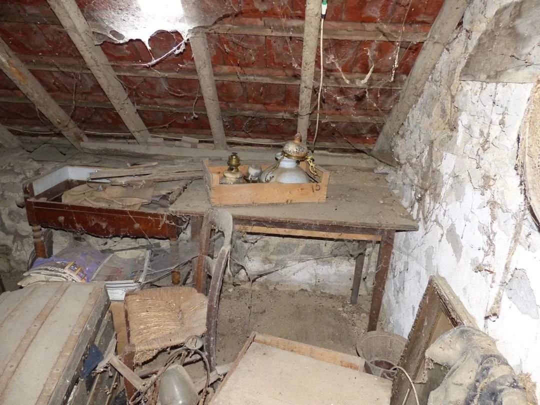 Old, dusty attic with dusty wooden furniture, a table, a chair, and various items covered in cobwebs and dirt, illuminated by natural light.