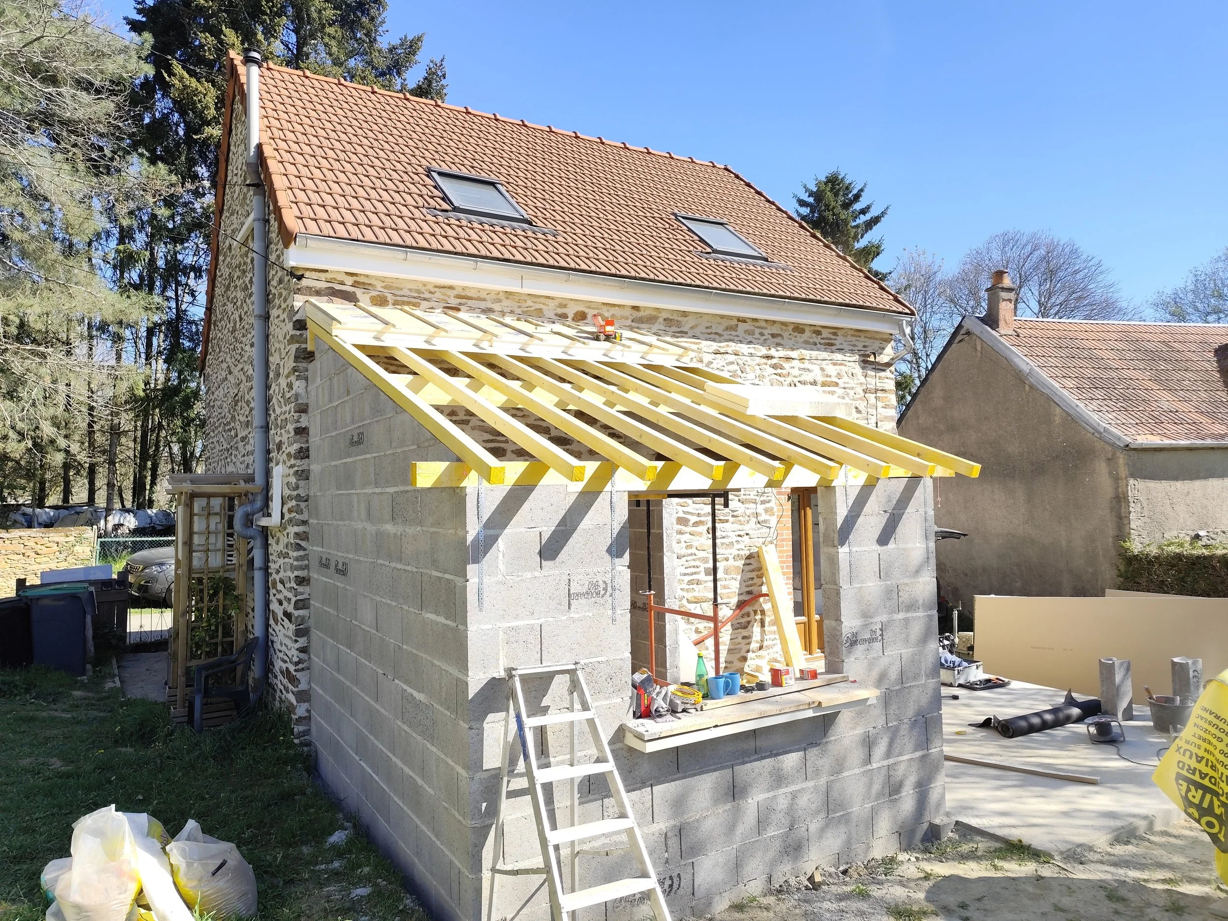 Construction site of a house with exterior walls built and roof partially under construction, scaffolding, and tools scattered around.