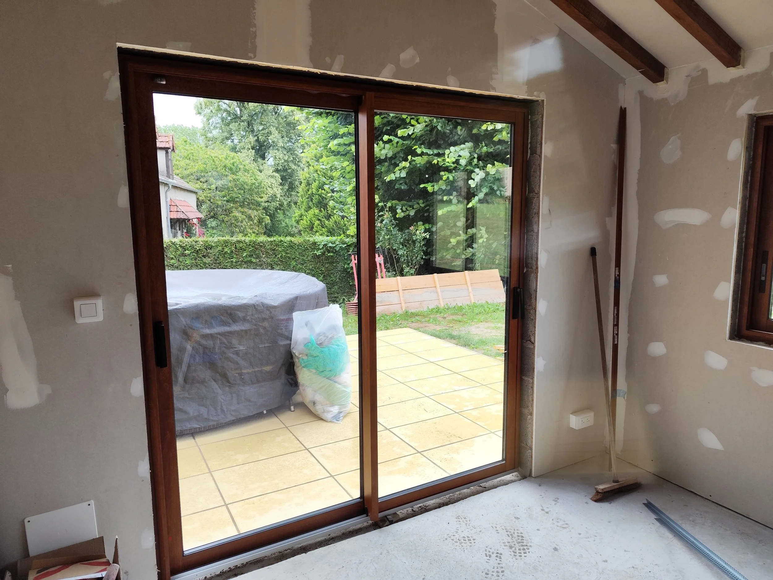 Inside a room under renovation with unfinished drywall on the walls, a sliding glass door leading to a patio, and construction materials and tools scattered around.
