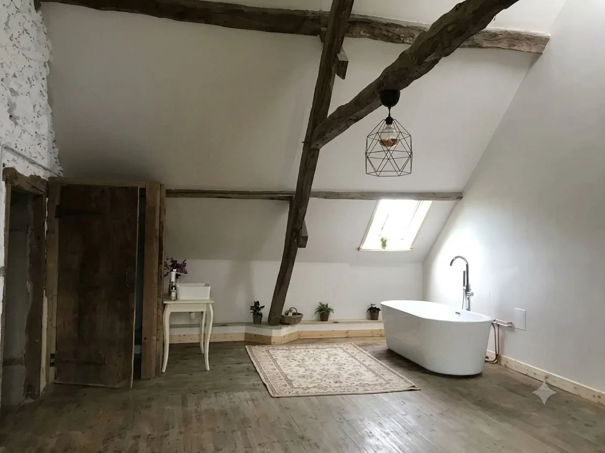 A rustic attic bathroom with a freestanding bathtub, small window, plants, a side table, and wooden beams.