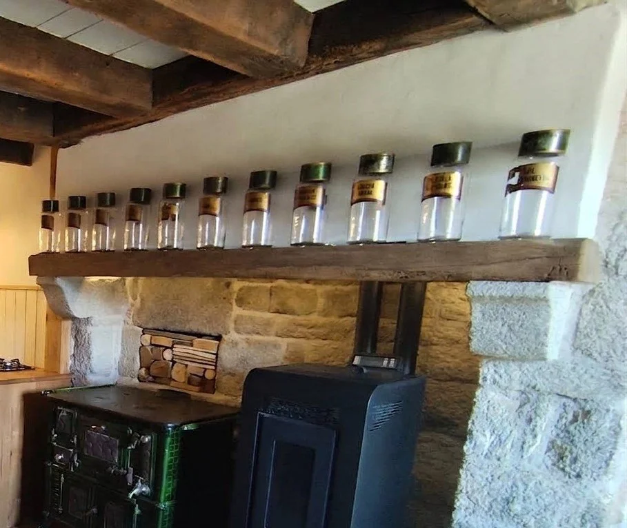 A rustic kitchen corner with a stone wall and wooden beams, featuring a wooden shelf holding empty spice jars with black lids and labels, and a black stove or oven below.