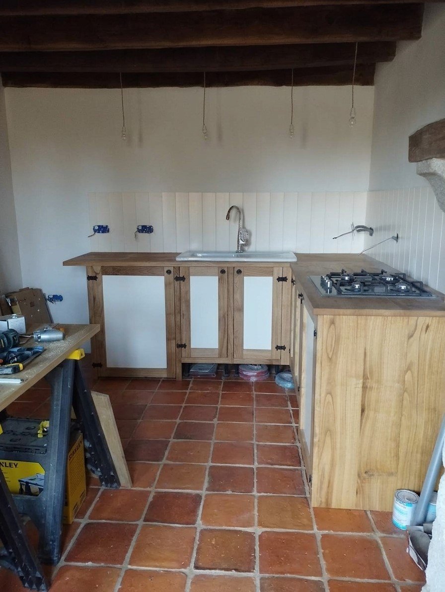 Partially built kitchen with wooden cabinets, a sink, and a gas stove, with hanging light bulbs and tools on a workbench.