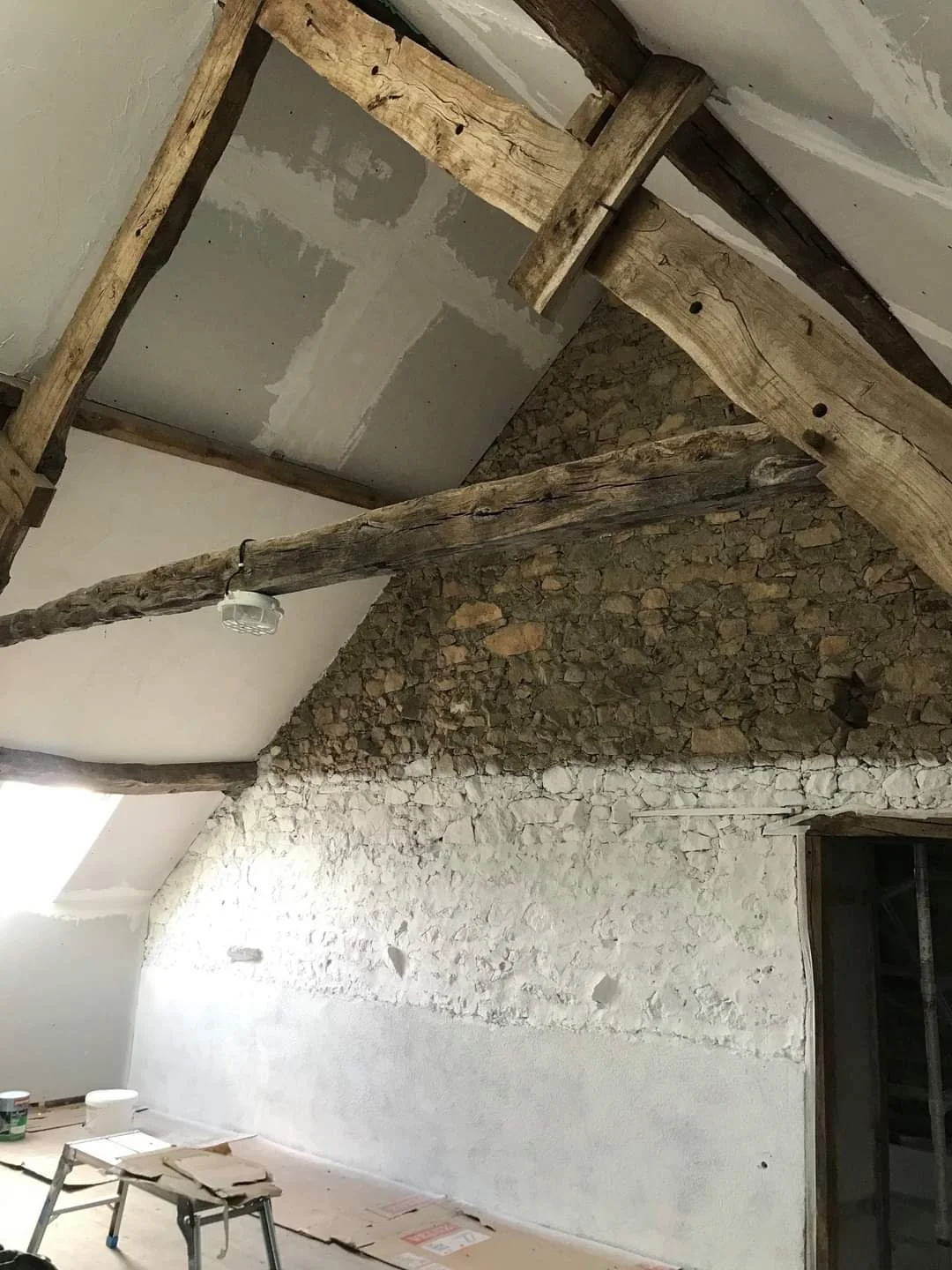 Interior of a room under renovation featuring an exposed stone wall, unfinished drywall, and rustic wooden beams on the ceiling.